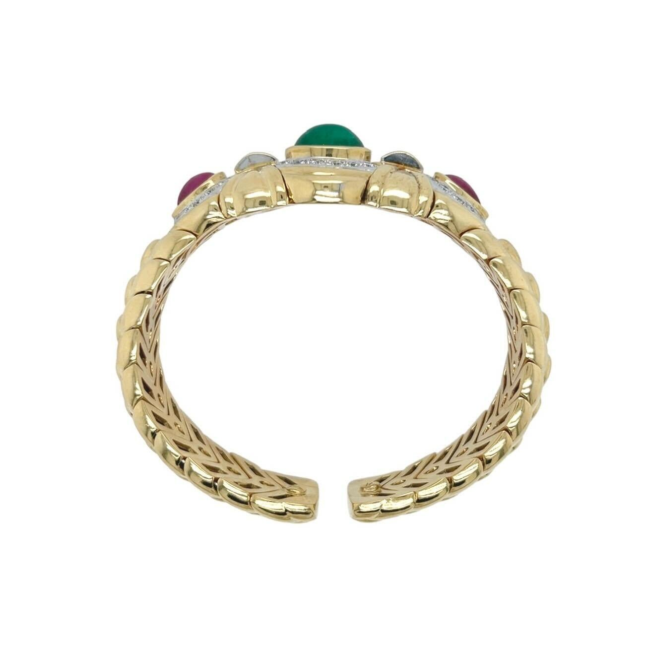 An 18 karat yellow gold, emerald, ruby and diamond bracelet.  Fashioned as an open backed flexible cuff of stylized braid design centering a bezel set oval cabochon emerald measuring approximately 10.85 x 7.58 mm surrounded by twelve (12) round