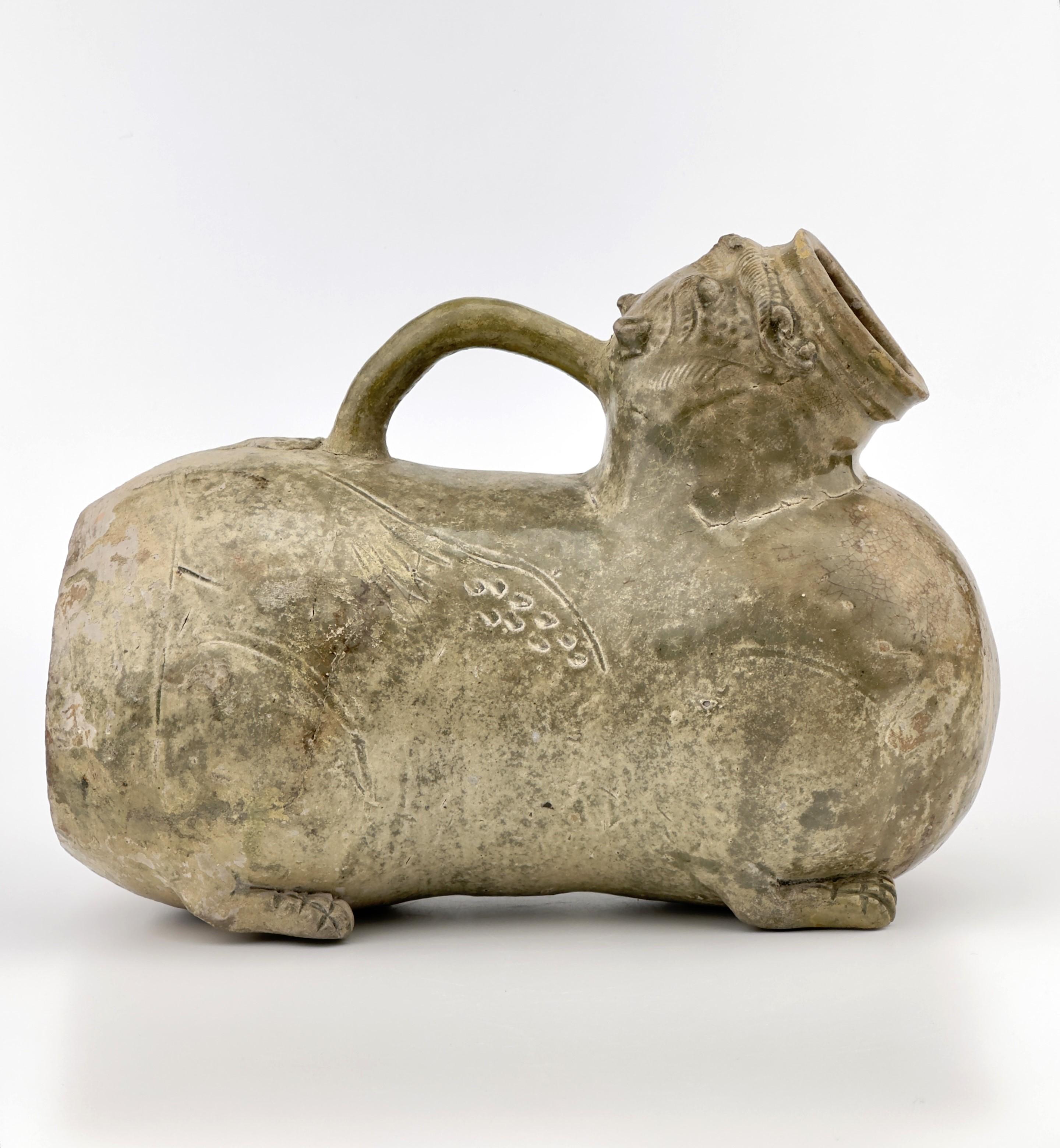 This vessel is well-modeled as a recumbent winged lion with detailed decorative elements. It features a grimacing face with large protruberant eyes under heavy brows and a gaping mouth, which forms a large aperture. The piece has a tail that arches