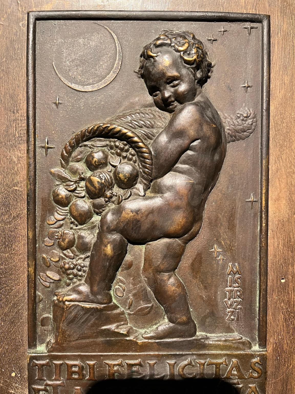 Plaque en bronze (dimensions sans le bois 12 x 20 cm) avec bas-relief représentant un putto avec une corne d'abondance et le croissant de lune. Dans la partie inférieure figure en lettres capitales l'inscription latine 