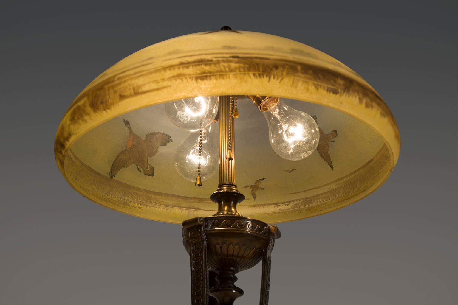 This is a beautiful brass and glass lamp with a flock of geese in flight over a water landscape. Shading of sky has the soft blue and golden color of an evening sunset. 

Pairpoint is known for three kinds of glass lampshades, originally produced