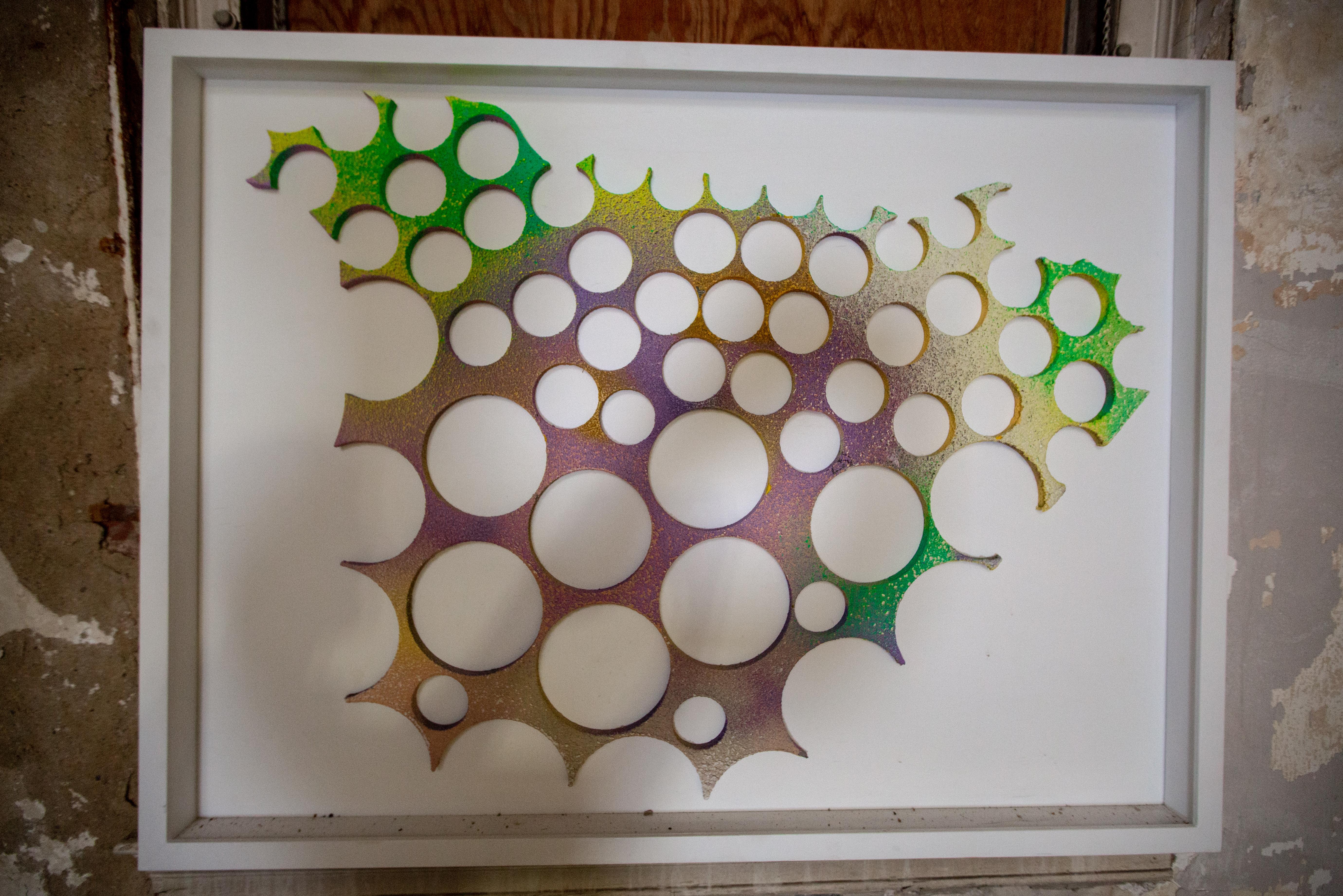 This unique piece by Joel Blenz has a white background with a white deep frame with a 3-d optical shape in the middle of it in many different colors. The piece has predominantly purple tones with green and yellow tones in the corners of the 3D