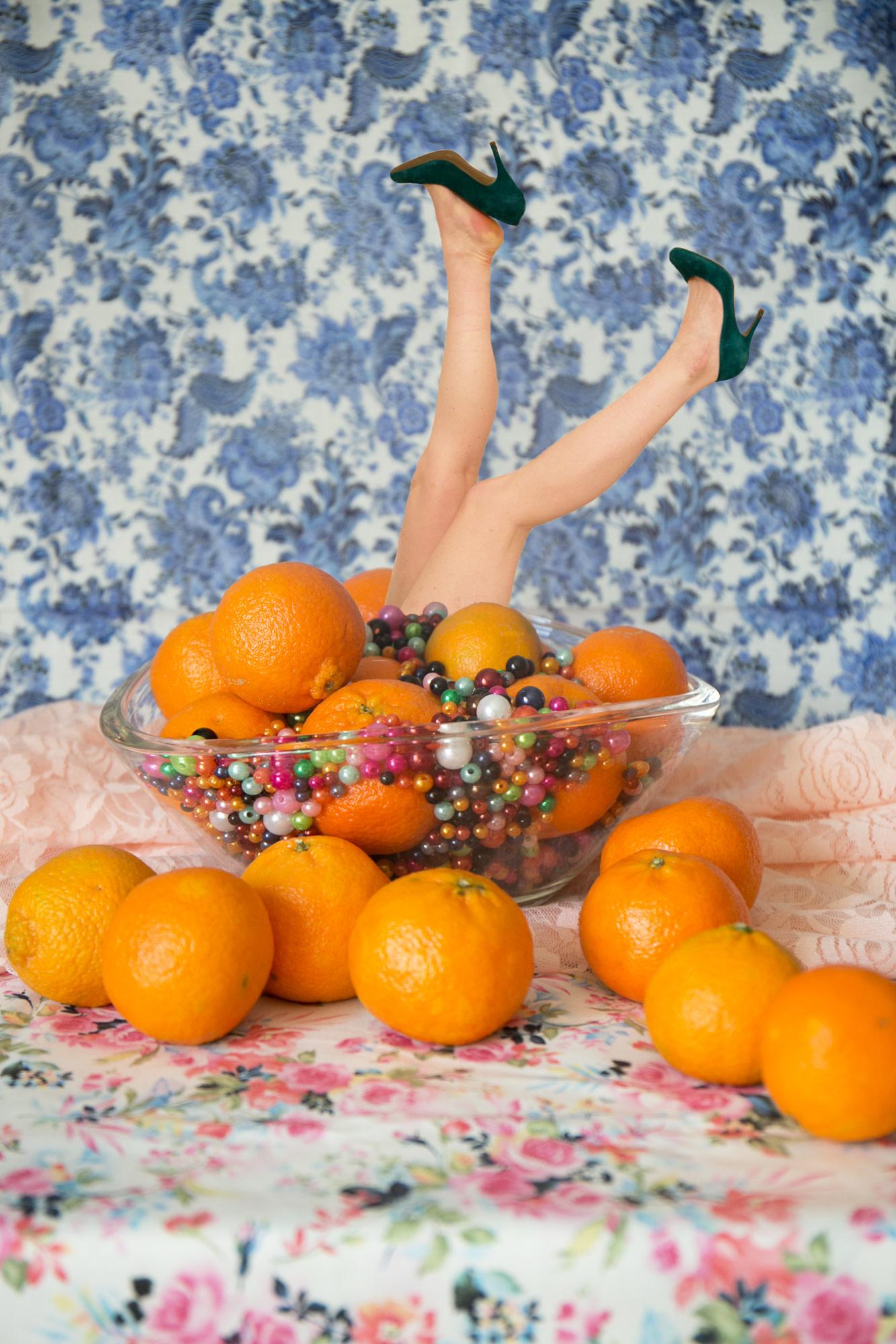 Julia McLaurin Color Photograph - Marge - Still life with oranges, a woman's legs, & floral wallpaper