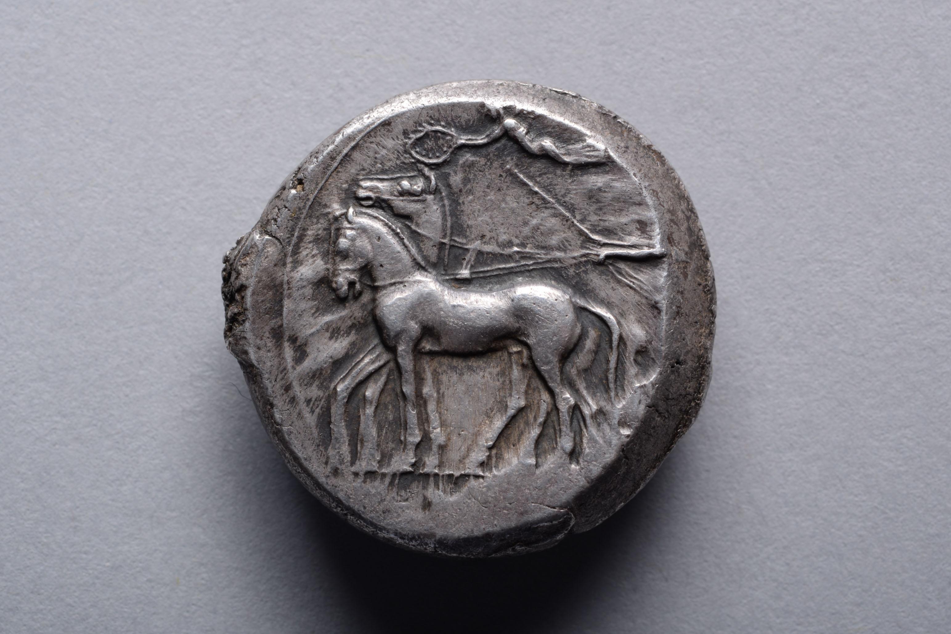 The obverse depicting a charioteer driving a four horse chariot, with the goddess of victory, Nike, descending from the heavens to crown the horses with a laurel wreath. The reverse with the image of the eponymous river god, Gelas, depicted in his