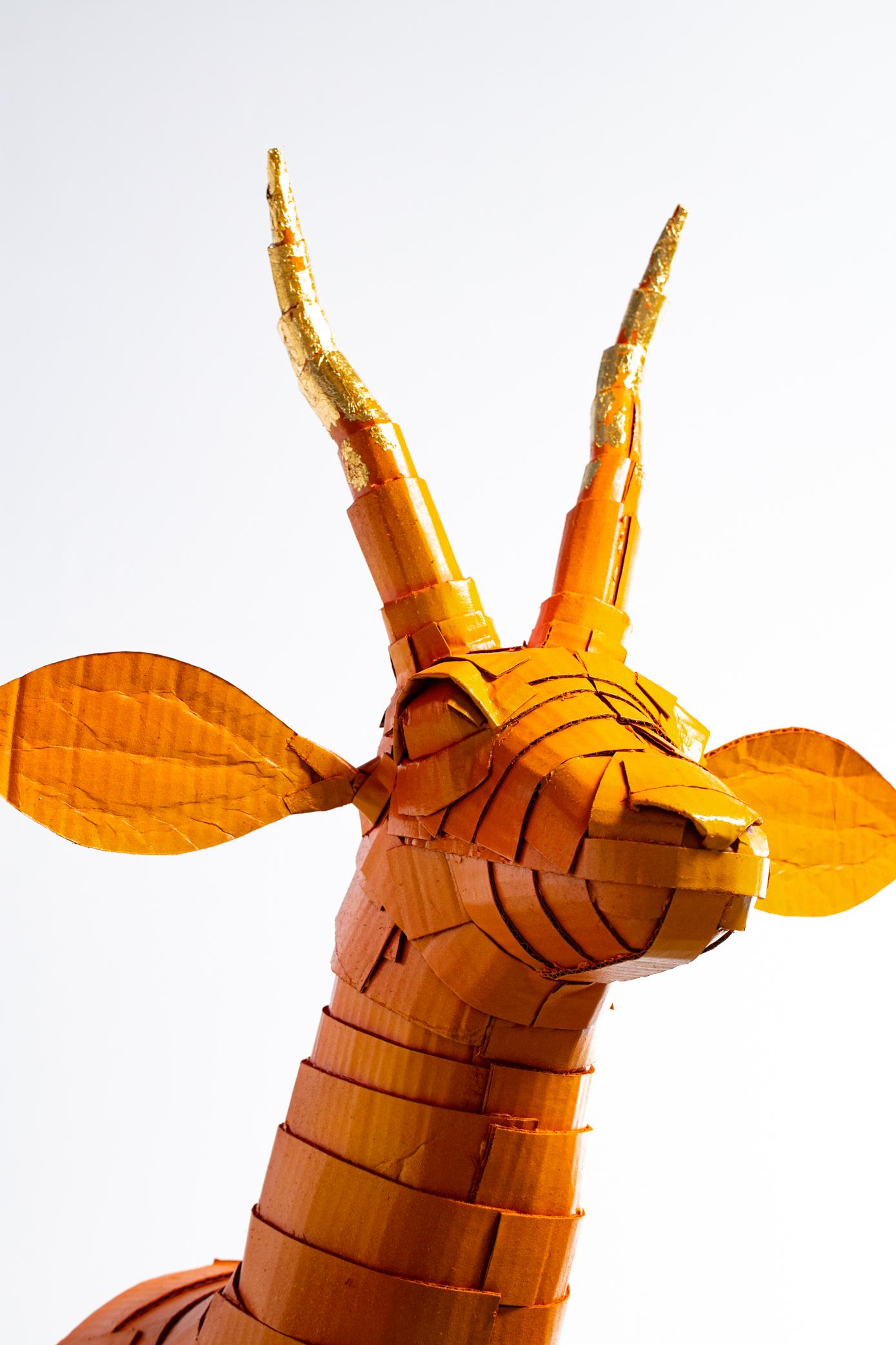 Sculpture de gazelle en orange kaki avec détail de corne en feuille d'or, méticuleusement créée par l'artiste Justin King à partir de morceaux de carton et de papier mâché découpés et pliés individuellement. Le choix des couleurs et les détails