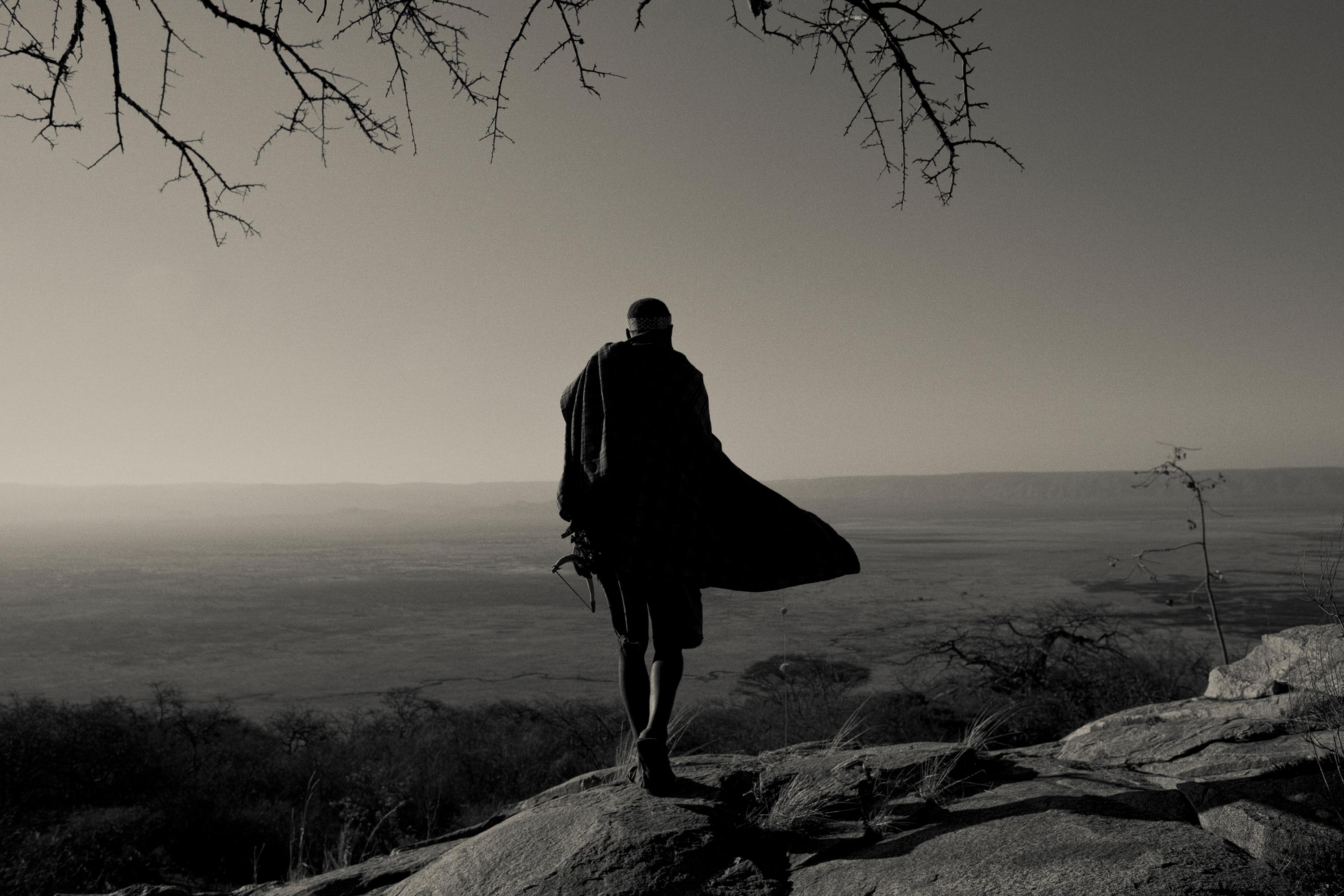 'HADZABE SKYLINE,'  a black and white photograph by Nicol Ragland of a tall tribal figure from the Hadzabe in Tanzania, overlooking the vast sparse landscape.  The archival giclee photographic print is 1/40 and is signed by the artist.  The print is