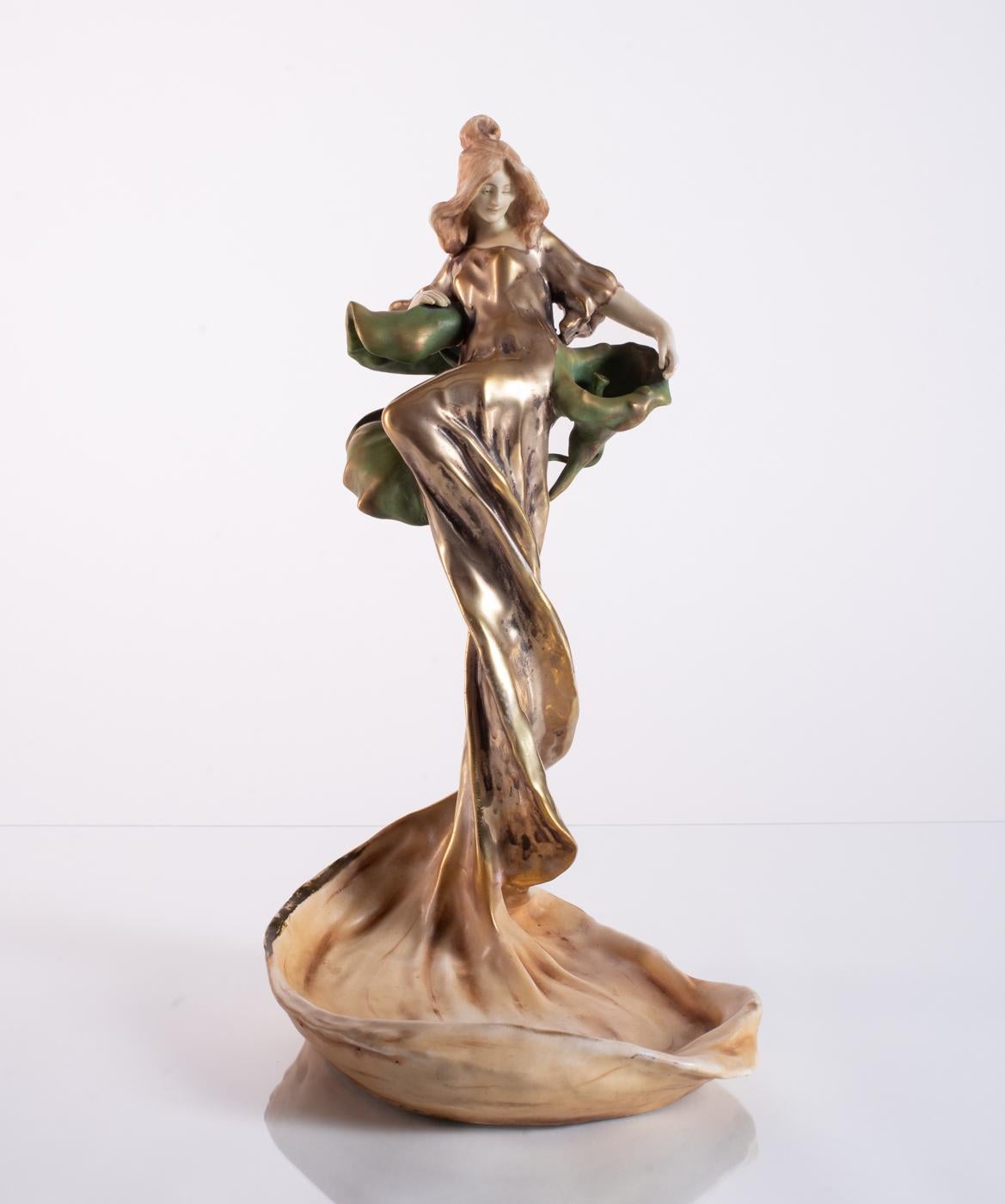 Incredibly dramatic Amphora ceramic featuring a maiden resting on a trio of lilies, her golden gown cascading into a gathered earthen base. Stamped Amphora in the base, and numbered.

Riessner, Stellmacher and Kessel (RStK), later known as Amphora,