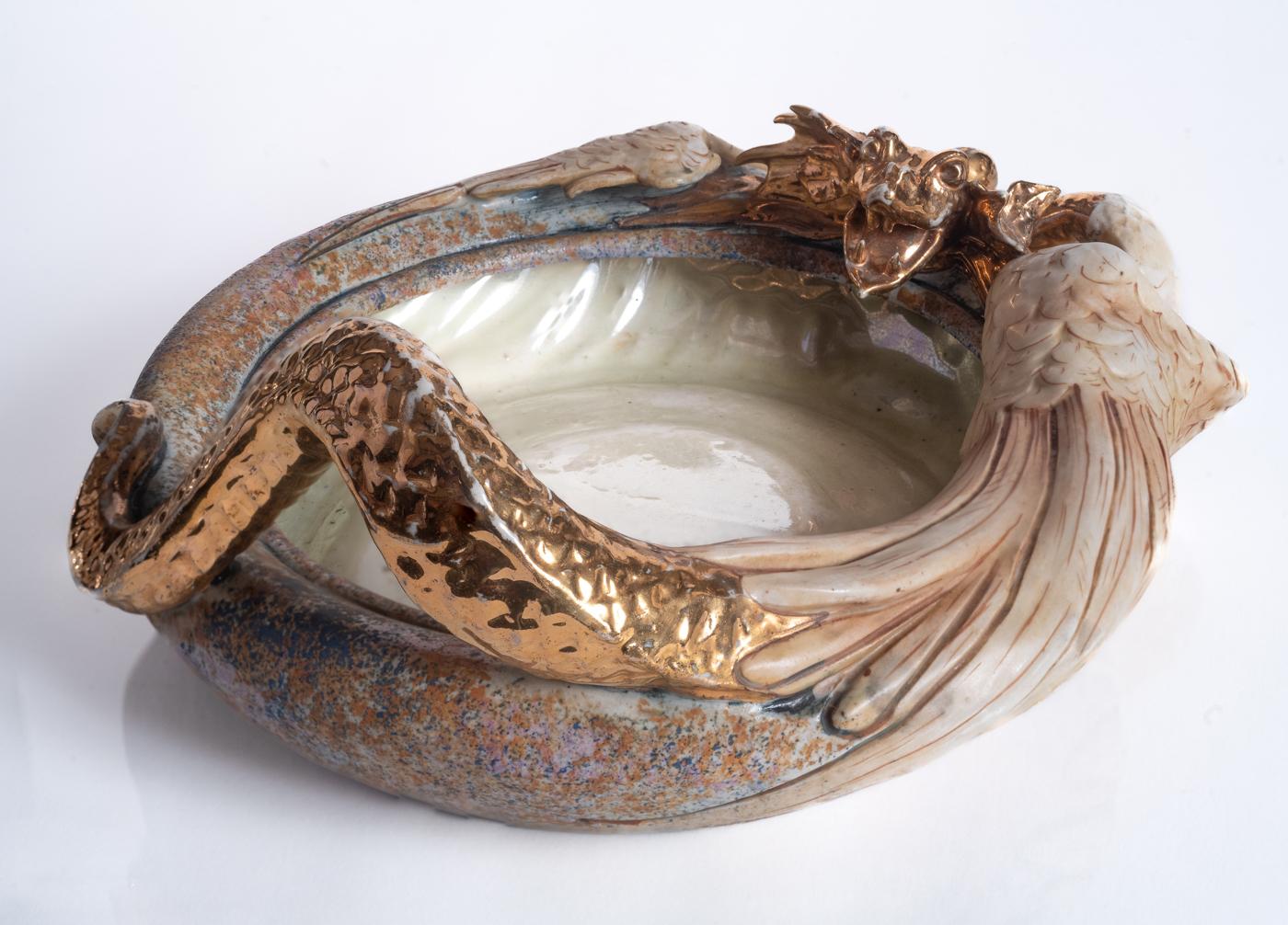 A golden serpentine dragon with feathered wings encircles a shallow bowl with mottled blue, pink, and orange glazing. Polished gold glaze on head and tail. This form was part of the collection selected for the 1900 Exposition Universelle (World’s