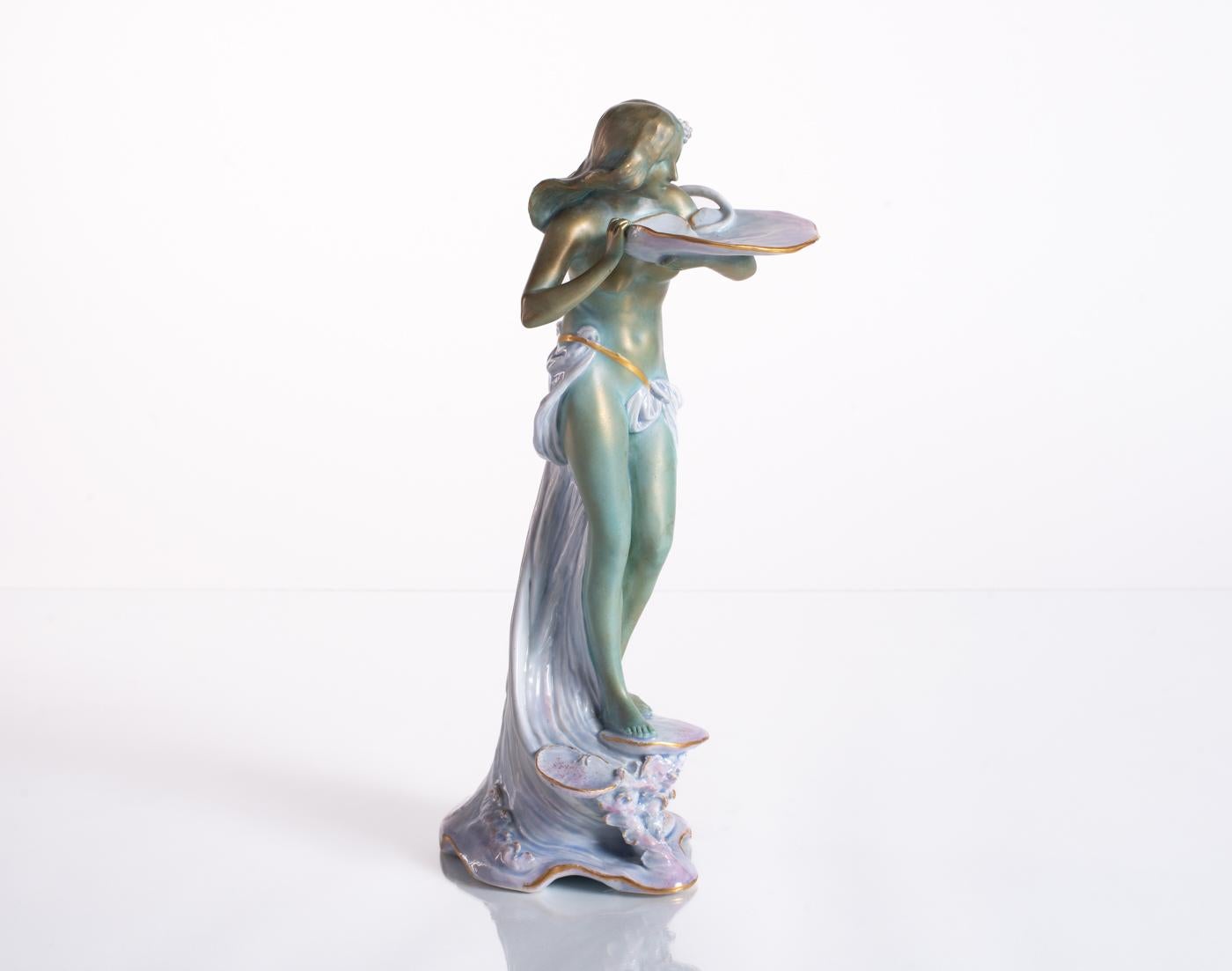 Standing atop a textured, frothy pastel sea, a classic green Art Nouveau maiden stands cradled by figural waves with an offering of a lily pad. An excellent example of the "ivory porcelain" ceramic material being used by producers in the