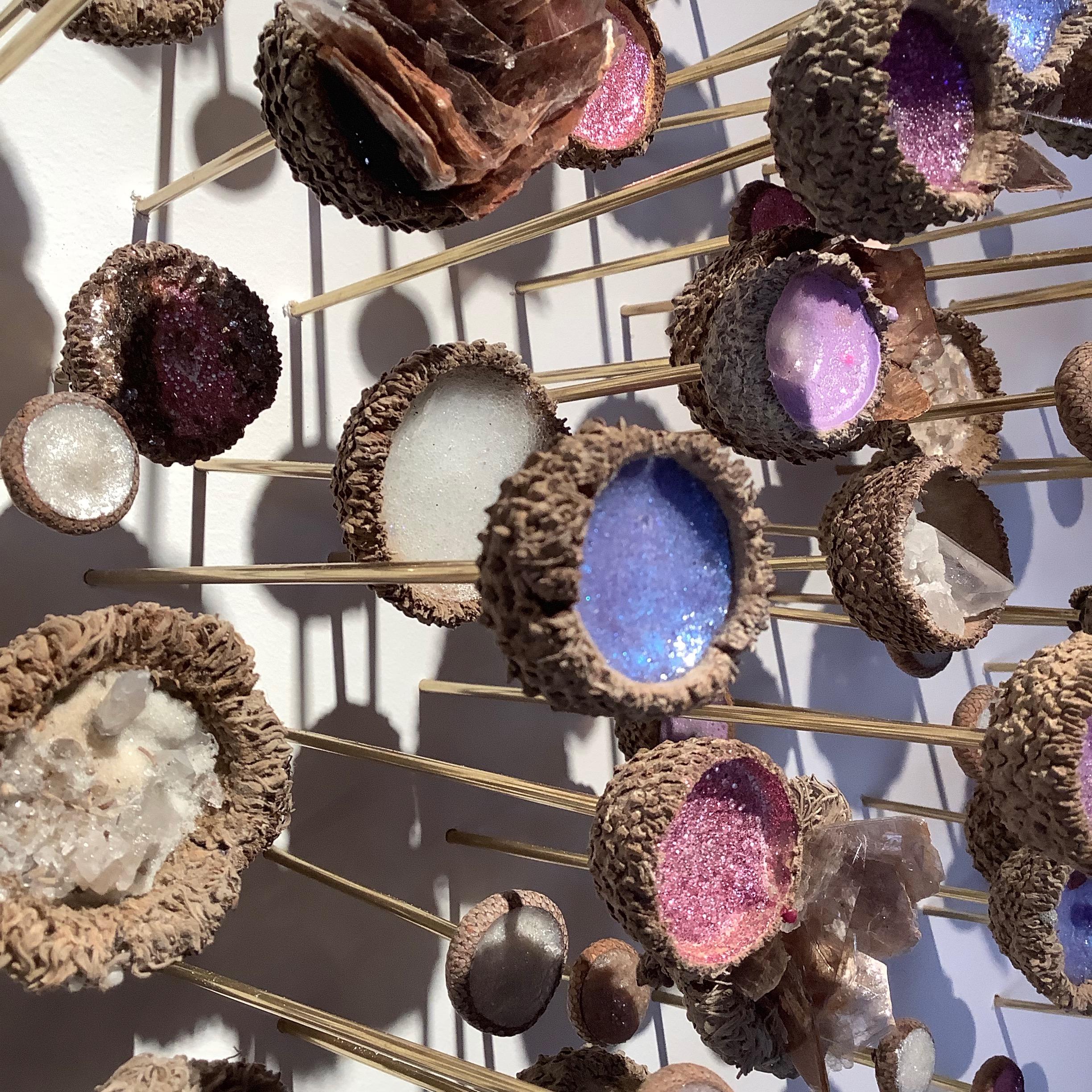 This wall sculpture consists of a meandering installation of acorn caps on brass pins filled with industrial pigments, glitter, natural minerals such as quartz and mica, and synthetic materials in hues of lavender, bright purple and pink, with