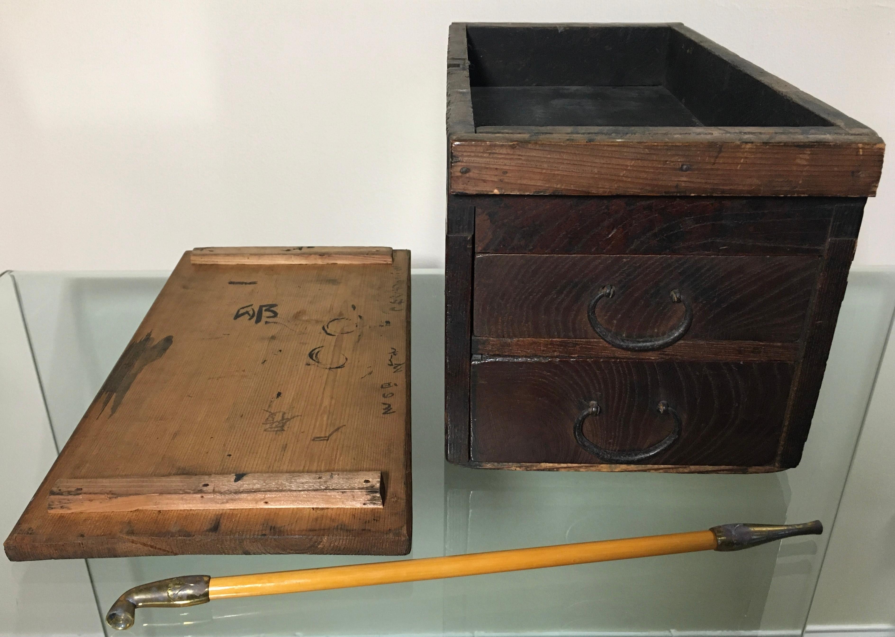Cette ancienne boîte à tabac japonaise, dotée de tiroirs, un kiseru, est une pièce d'histoire captivante. La torsion chaude de l'orme raconte une histoire de temps.
La boîte présente des accessoires métalliques authentiques qui racontent le riche