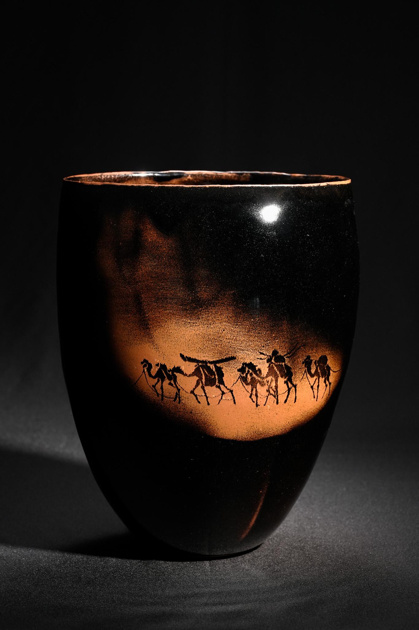 The Iron-Glazed Camel Vase by Yasuhisa Nagira is a powerful fusion of form, texture, and symbolism. Cloaked in a deep iron-rust glaze, the vessel exudes a dark, lustrous sheen that shifts subtly between brown and black, evoking both the richness of