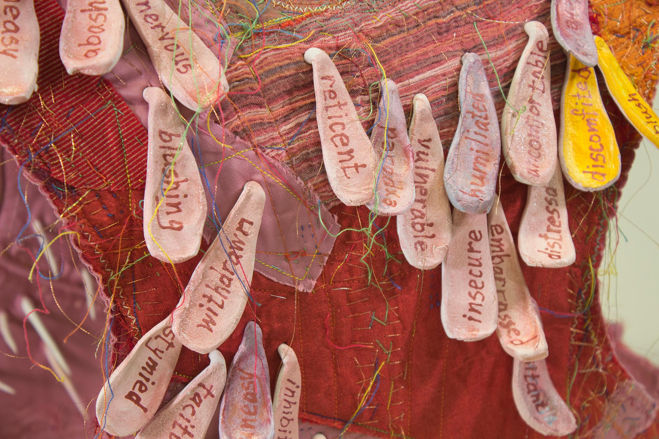 Virginia Mahoney’s “Blush” is a mixed-media sculpture in vest form that is pink, red, and orange, with attached ceramic tags on which words referring to embarrassment are handwritten. Ceramic spikes projecting inside emphasize discomfort, and