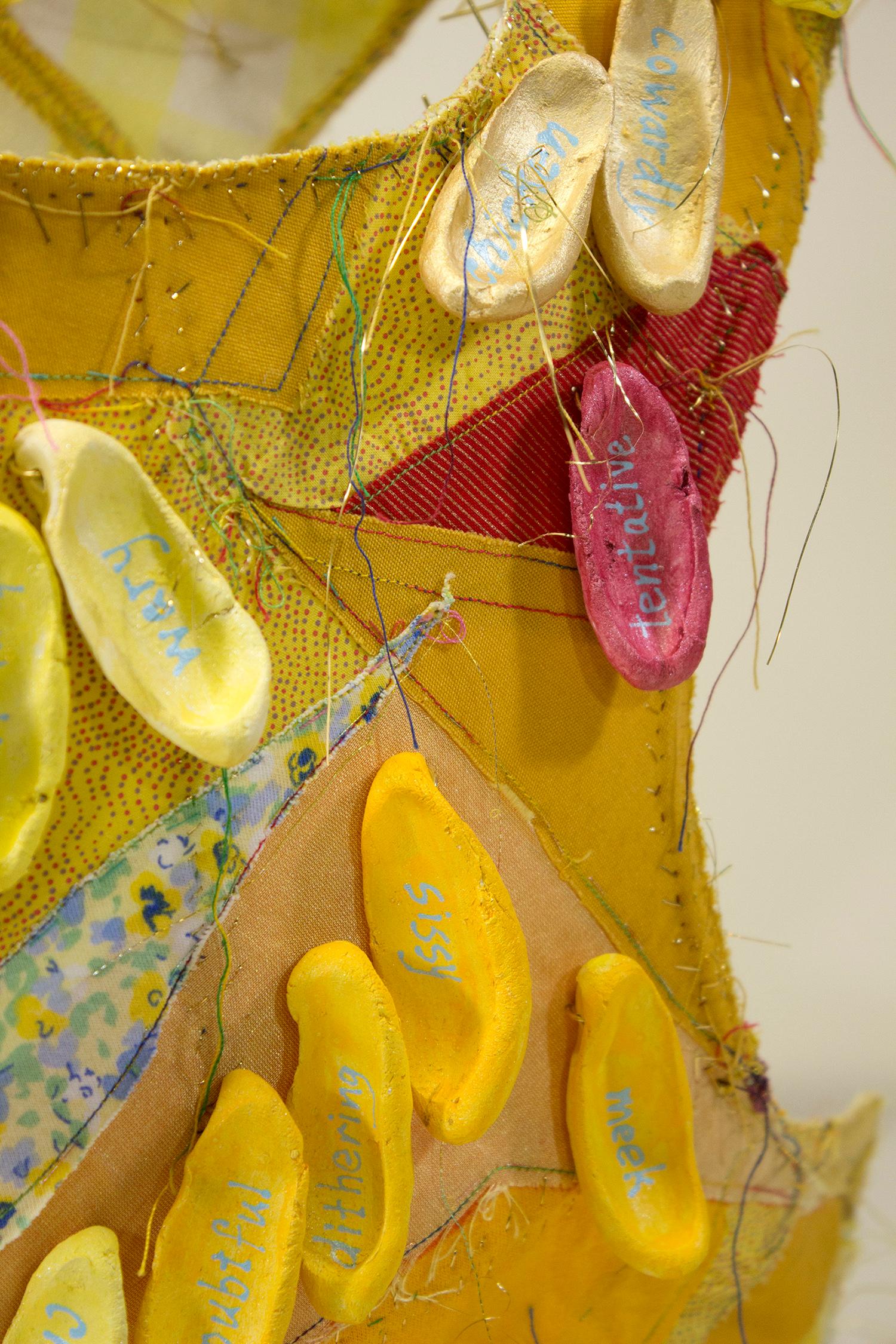Virginia Mahoney’s “Yellow” is a mixed-media sculpture in vest form that is yellow with red accents, and has attached ceramic tags on which words referring to fear are handwritten. Brass spikes projecting inside emphasize the discomfort one feels