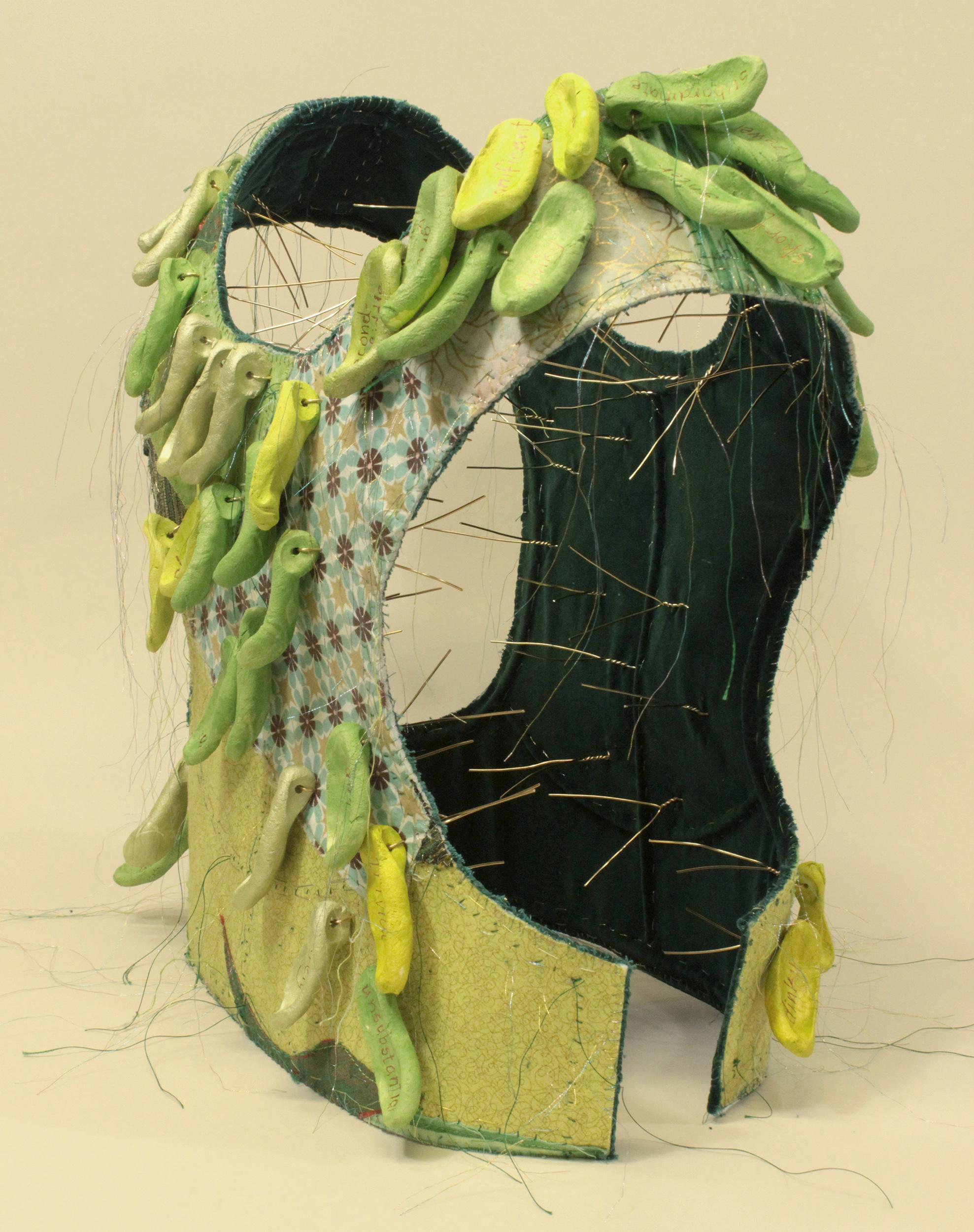 Virginia Mahoney’s “Less” is a mixed-media sculpture in vest form that is primarily green with many prints and variations, and has attached ceramic tags in shades of green, on which words, referring to being less than others, are handwritten in