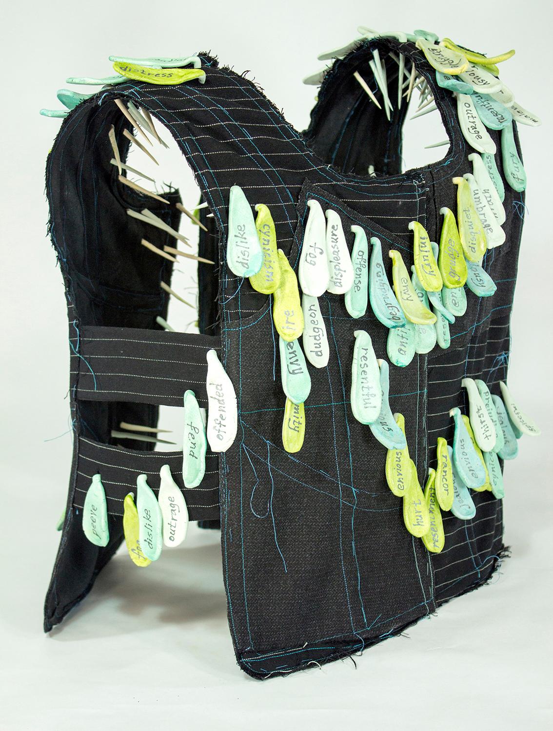 Virginia Mahoney’s “Envy” is a mixed-media sculpture in vest form that is black with white pinstripes, and has attached ceramic tags in various shades of green, on which words referring to being envious are handwritten. Ceramic spikes projecting