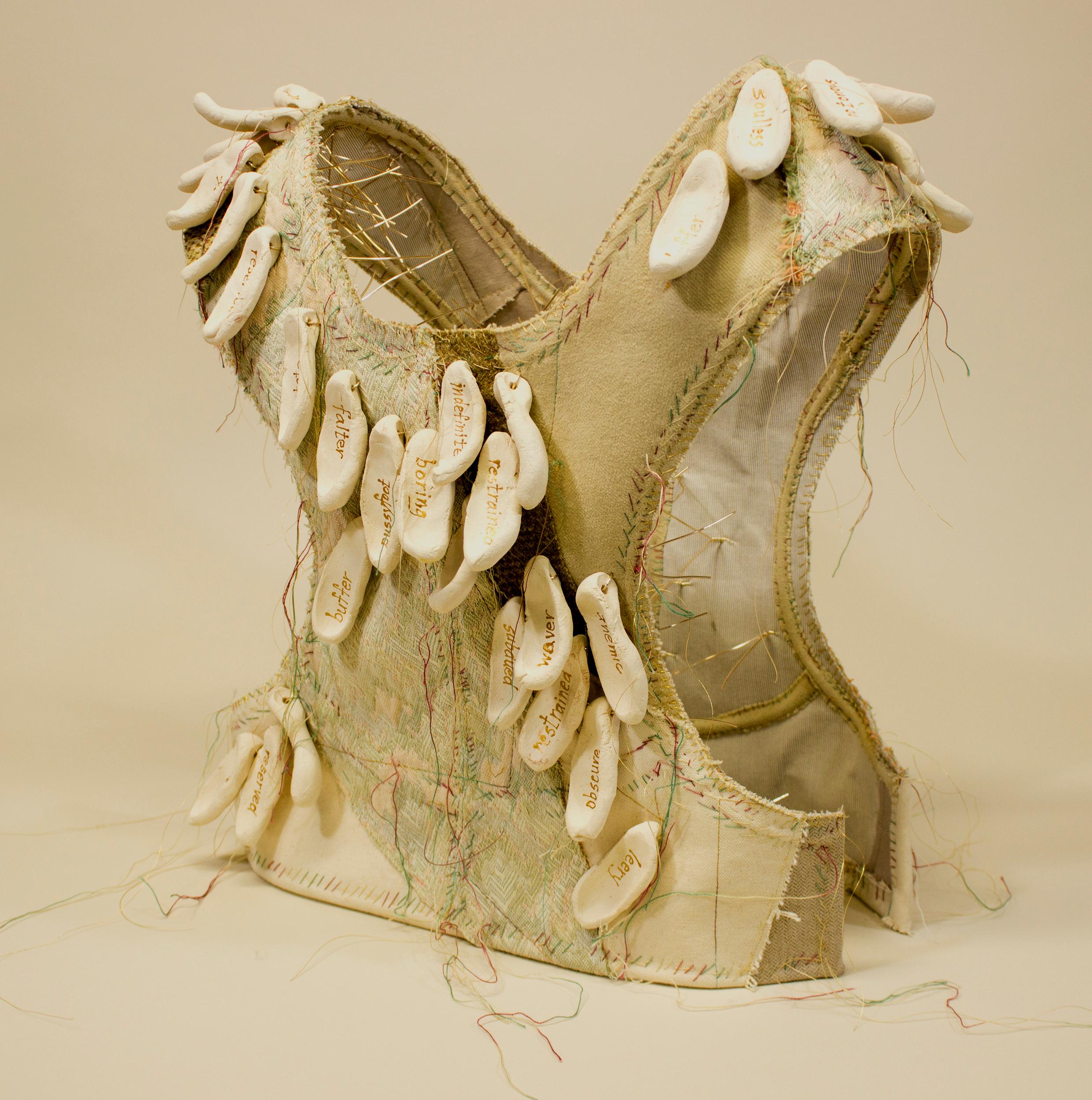 Virginia Mahoney’s “Noncommittal” is a mixed-media sculpture in vest form that is neutral in overall color, with several textures and variations. It has attached ceramic tags in off-white, on which words, referring to being unable to make decisions