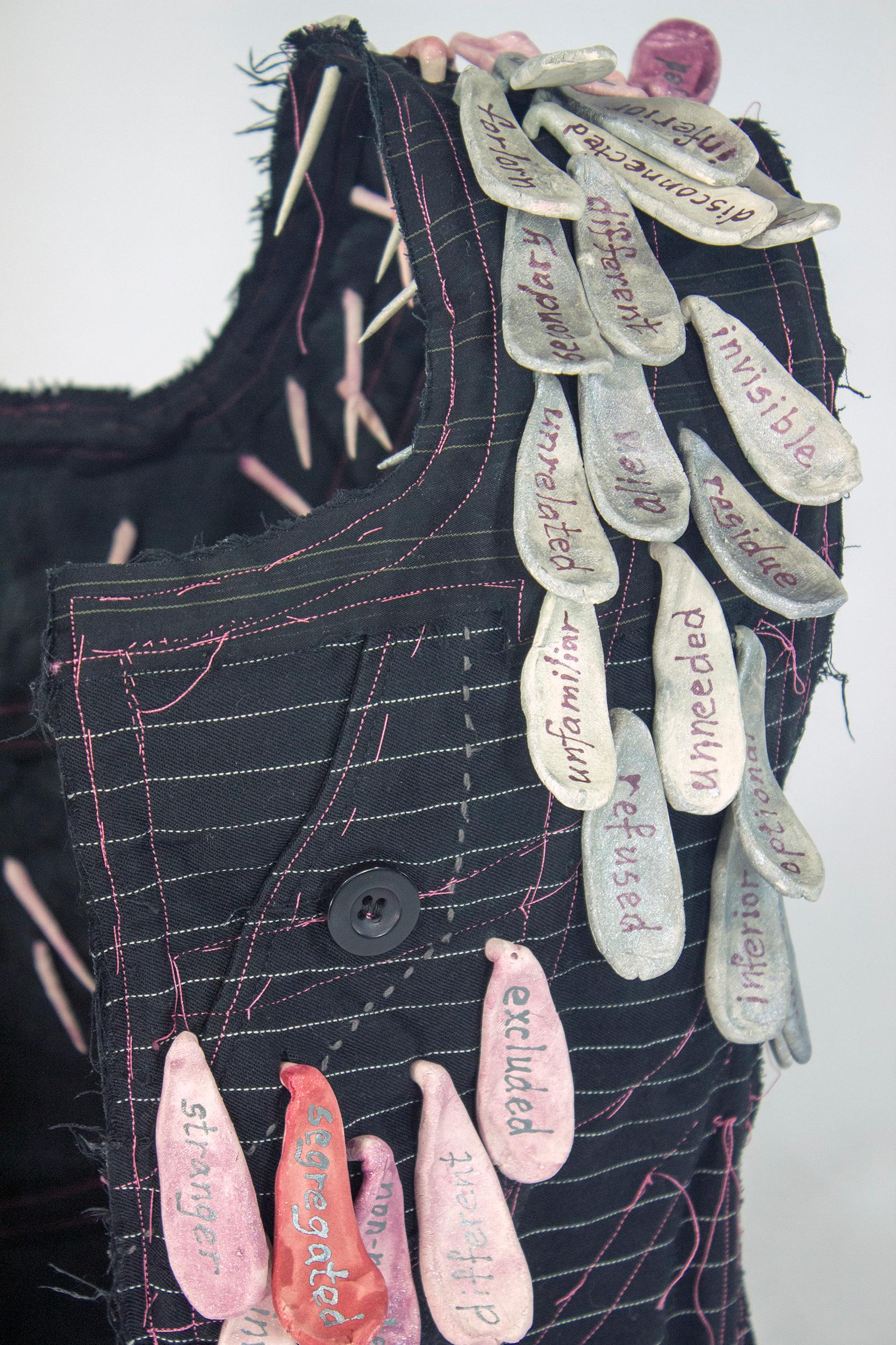 Virginia Mahoney’s “Other” is a mixed-media sculpture in vest form that is black with white and grey pinstripes, and has attached ceramic tags in various shades of pink, on which words referring to being ostracized or not included are handwritten.