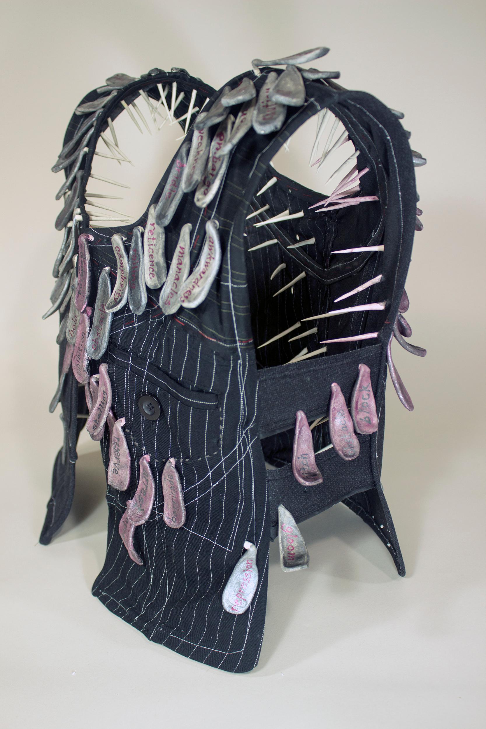 Virginia Mahoney’s “Restraint” is a mixed-media sculpture in vest form that is black with light pinstripes, and has attached ceramic tags in various shades of grey or muted pink, on which words, referring to practicing restraint, are handwritten in