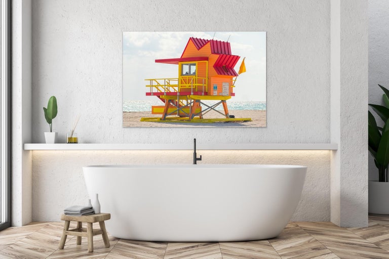 This contemporary coastal photograph by Peter Mendelson features a signature Miami Beach lifeguard stand at the center. The bright orange structure features yellow railings and a red, star-shaped roof. The lifeguard stand is situated in the sand,