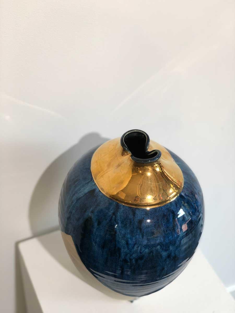 This modern ceramic vessel by Connecticut-based ceramicist, Jon Puzzuoli, is made with glazed porcelain. A brilliant blue glaze is dripped down the top half of a textured white base, and the mouth of the vessel is finished with 18k gold luster.