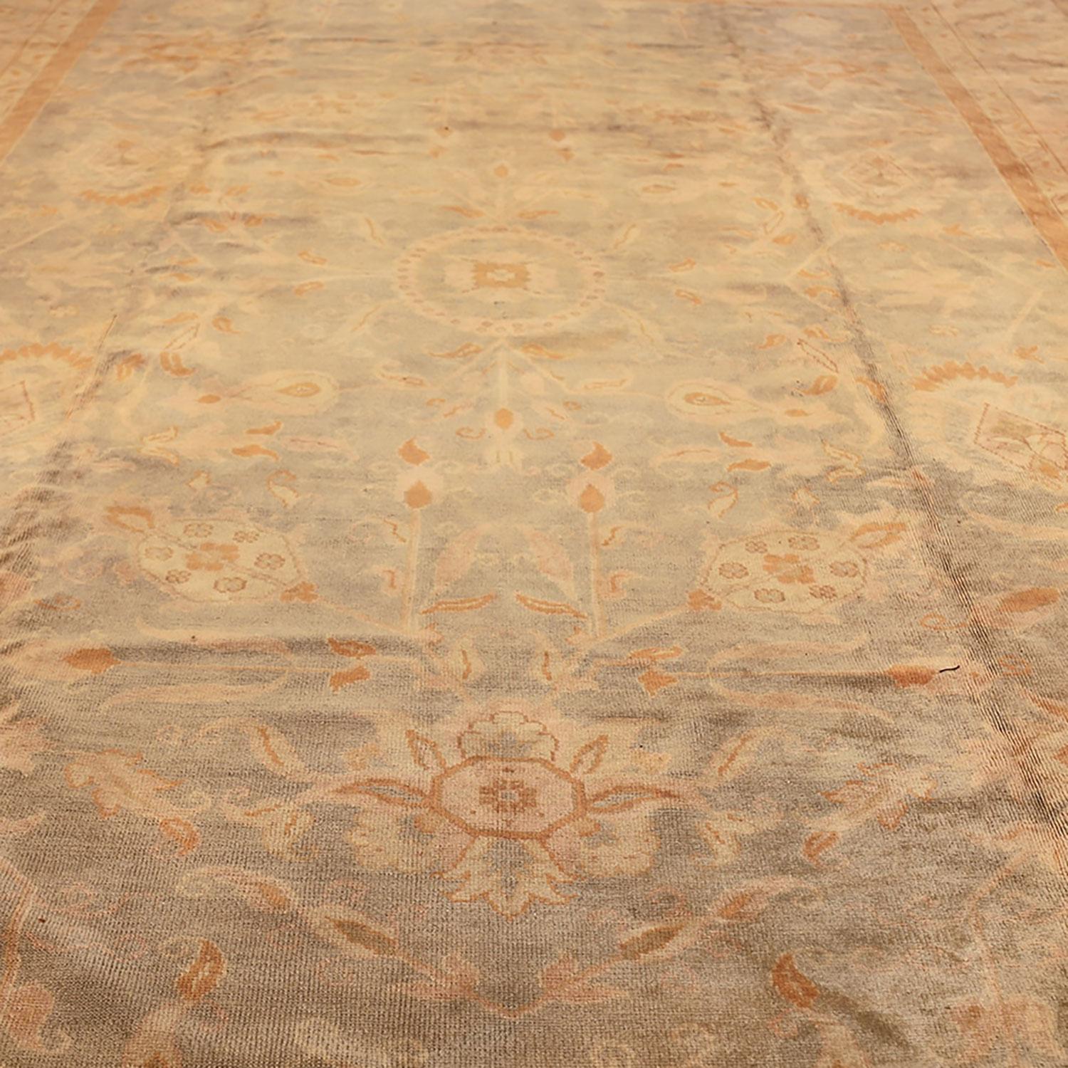 This antique Turkish Oushak wool rug draws you in with its warm, honeyed glow, a timeless testament to the early 1900s. Crafted with elegance and handwoven in Turkey, this 11’ x 15’ rug features an intricate floral motif anchored by a delicate,