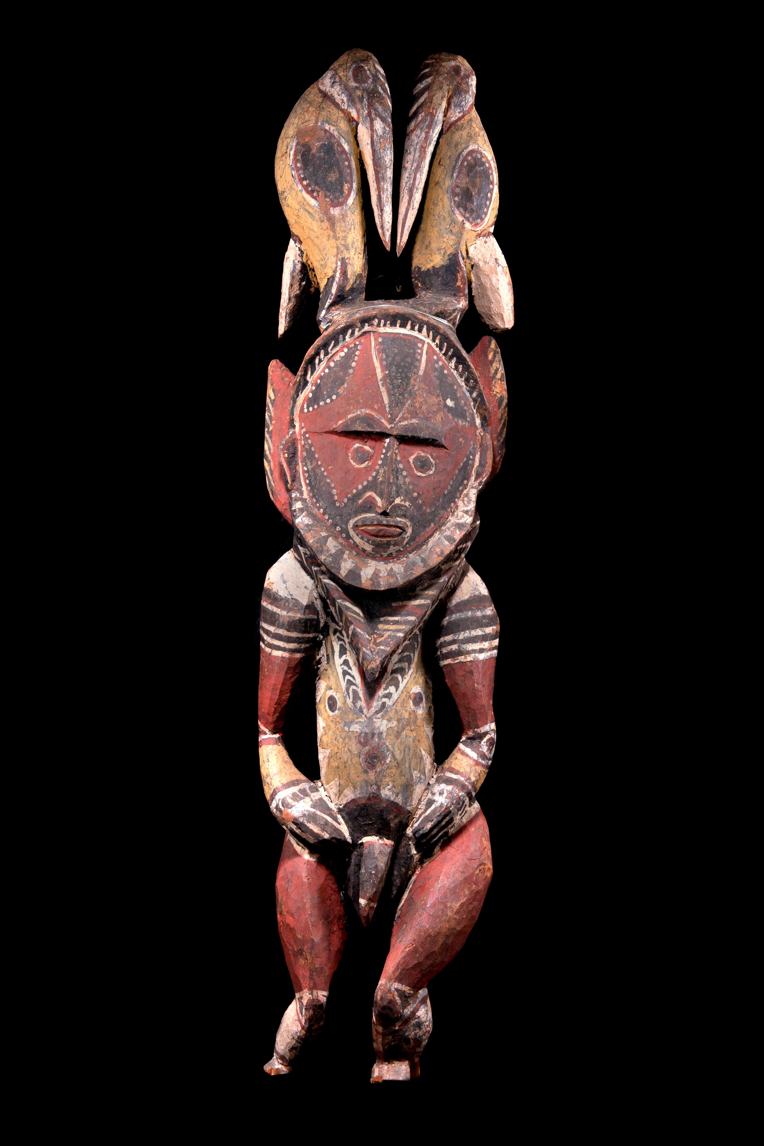Full male figure standing with legs bent inward, pointed penis, arms carved away from body and hands resting at hips. The large oval face framed by beard, extensions behind ears, and surmounted by pair of tall hornbill birds. Front of surface