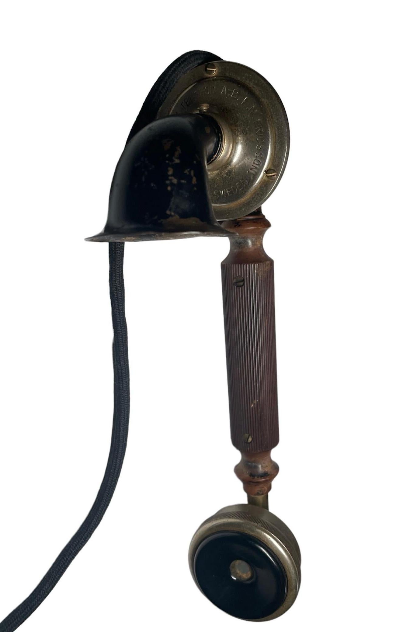This is a A.B.L.M. Ericsson Sweden hand crank table/desk telephone as it is hallmarked in the handset. It is a rectangular heavy metal box painted black. The metal box is supported by four round metal bottom shaped feet. The handset is made of wood