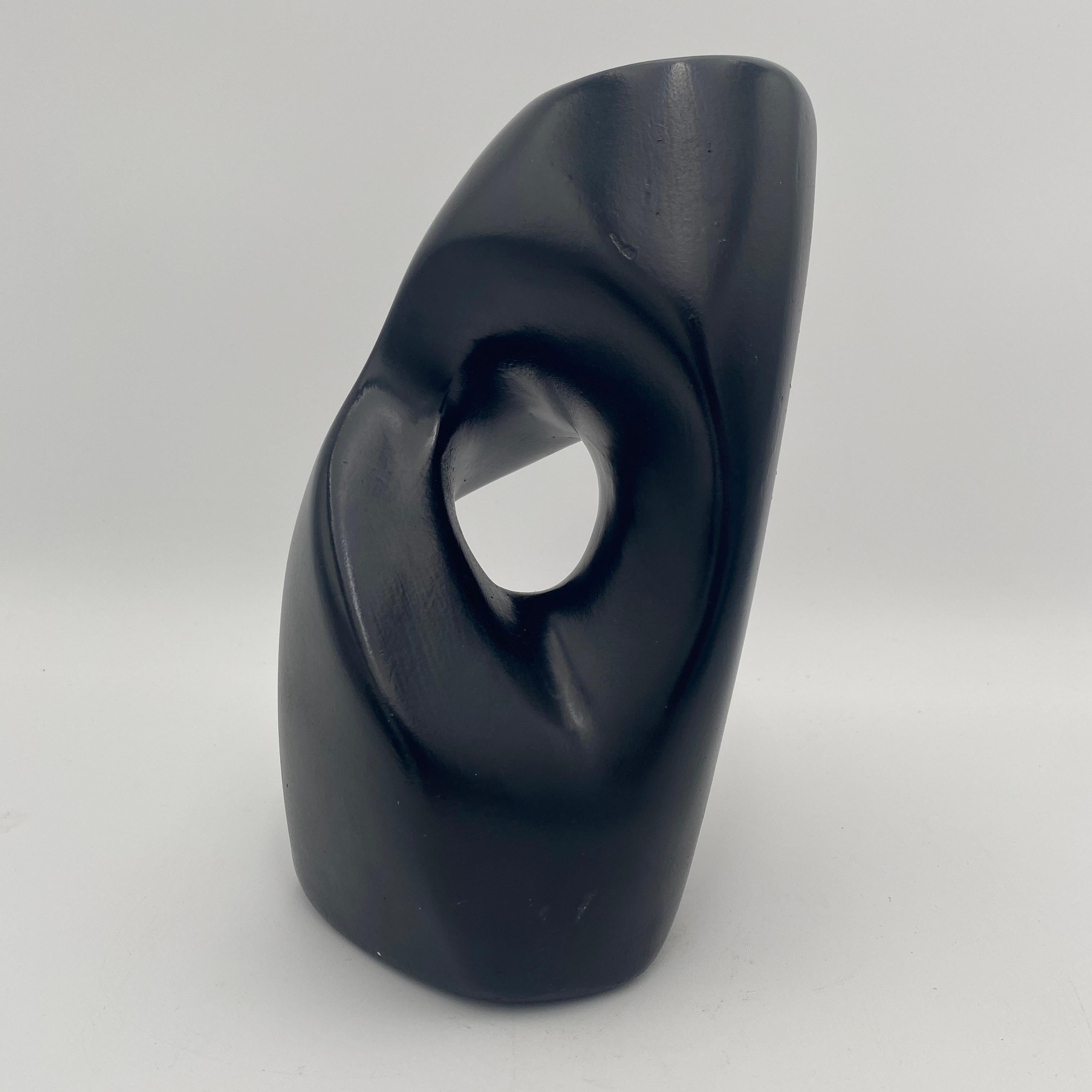 This abstract sculpture has a smooth black, glossy surface and an organic modernist form. The sculpture has a negative space that pierces through its center creating a sense of depth and complexity. The shape is fluid and curved, resembling a