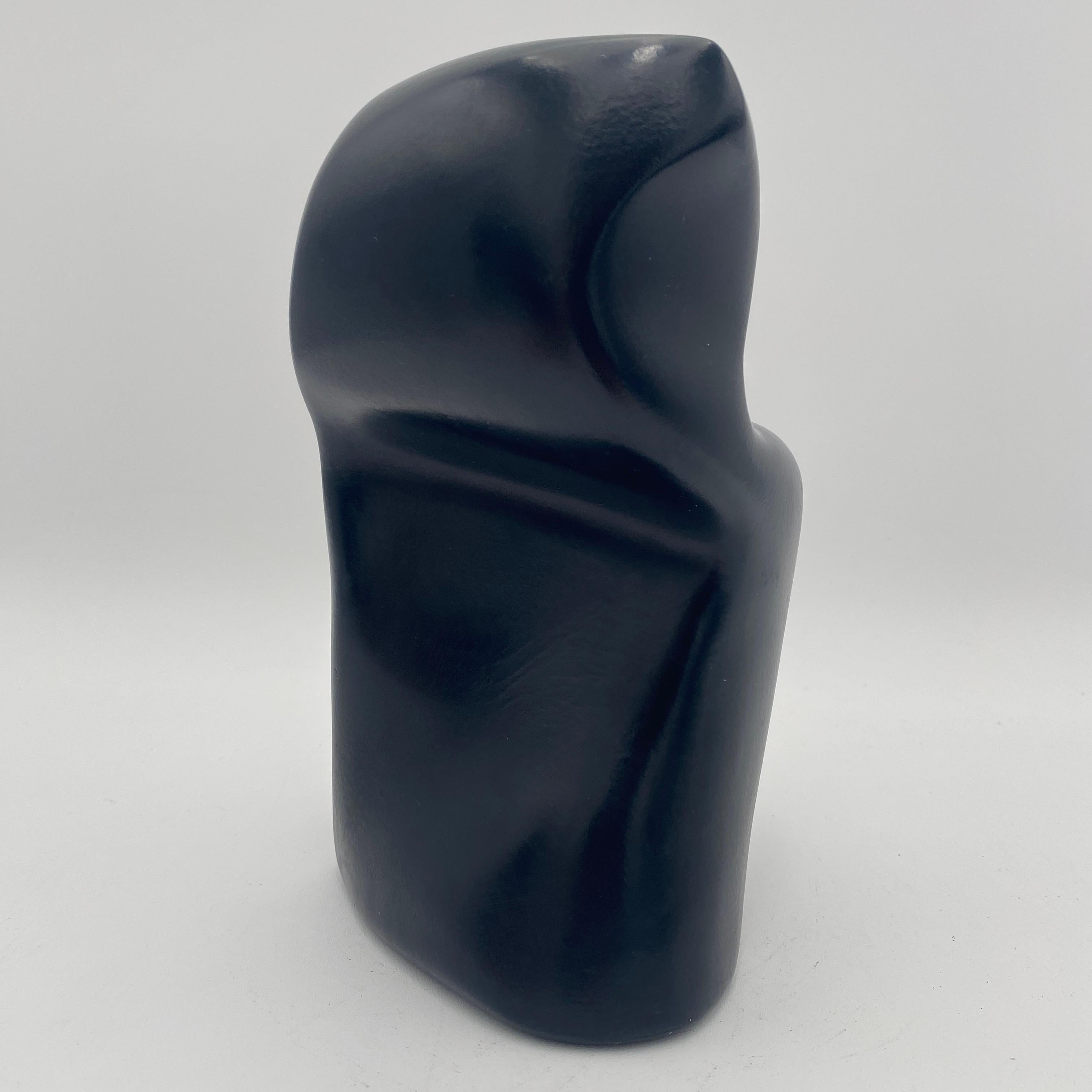 This abstract sculpture has a smooth black, glossy surface and an organic modernist form. The shape is fluid and curved, resembling a stylized human form or a natural element. The dark color and smooth texture give it a modern and elegant