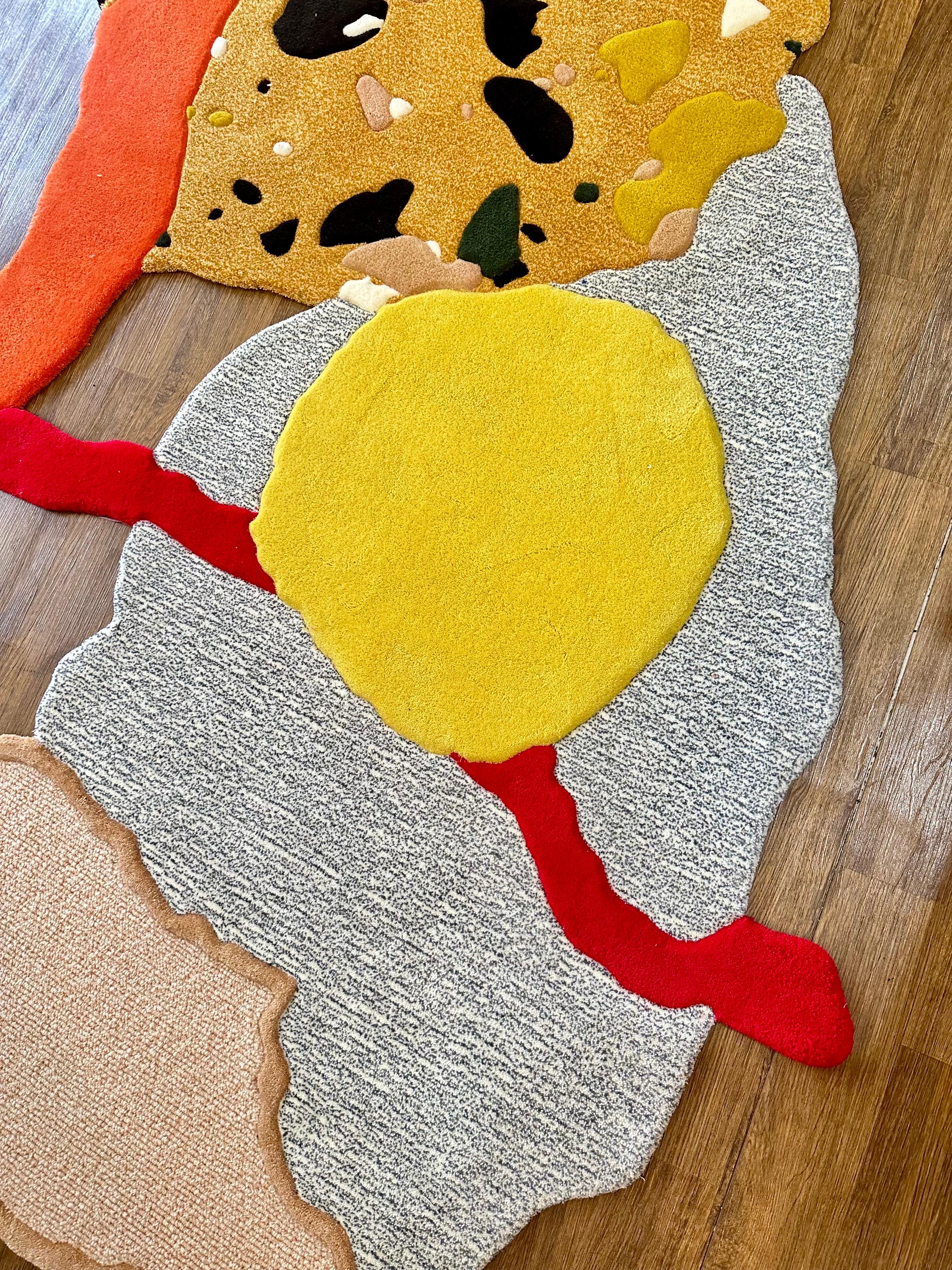 Dieser kühne, moderne Teppich kombiniert eine von Terrazzo inspirierte Musterung mit lebhaften abstrakten Formen in Rot, Gelb, Orange, Beige und Grau. Die organische Silhouette unterstreicht seine skulpturale Präsenz und lässt ihn eher wie ein