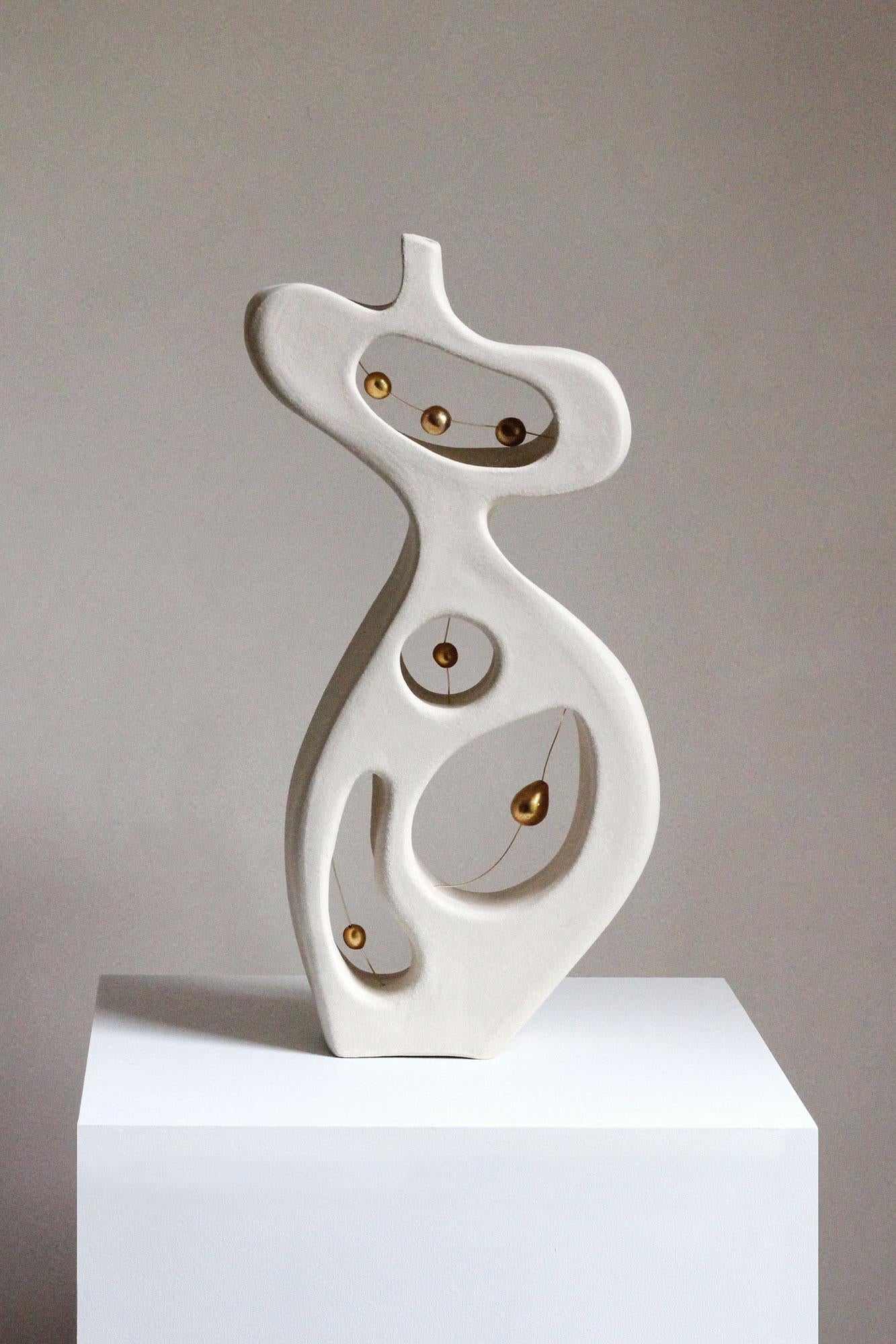 Réalisée à la main en grès, cette sculpture abstraite incarne la forme galbée d'une femme. Les creux de l'espace négatif à l'intérieur du corps permettent de voir à travers l'œuvre, qui présente des fragments d'or 24 carats qui flottent, suspendus à