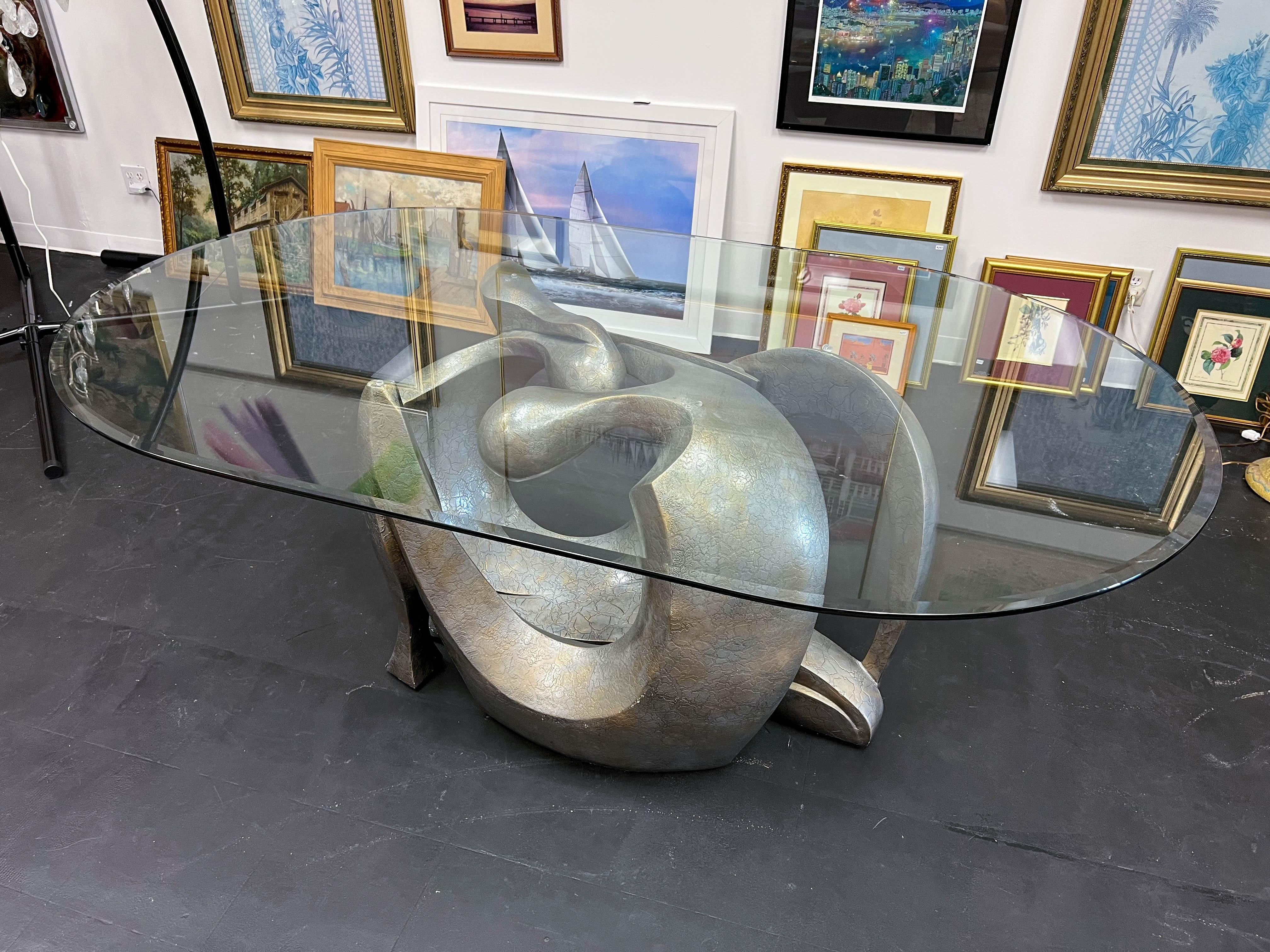 The abstract table base consists of two intertwined figures, evoking the union between male and female. Textured and finished in silver with delicate hints of gold, the base fabricated of composite is topped with an oval-shaped beveled glass. The