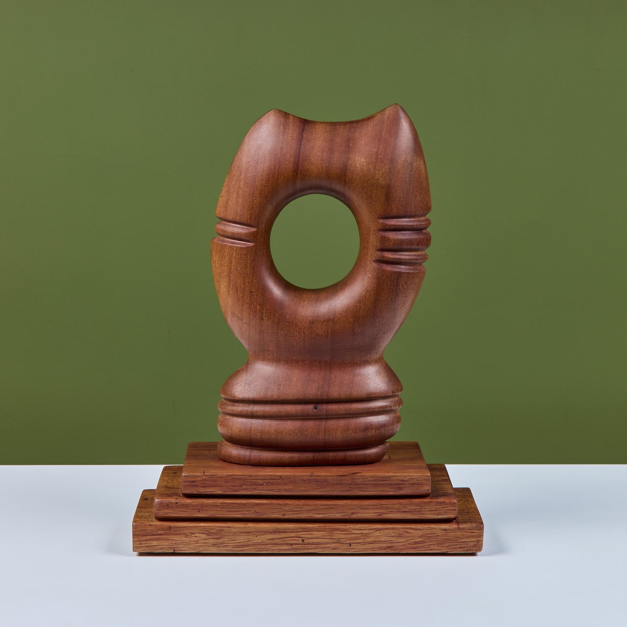 Hand-carved wooden abstract cat head. The piece showcases two pointed ears and whiskers on each side of the face. The center of the sculpture has a round hole at the middle and is mounted to a three tier stacked base. Signed by the