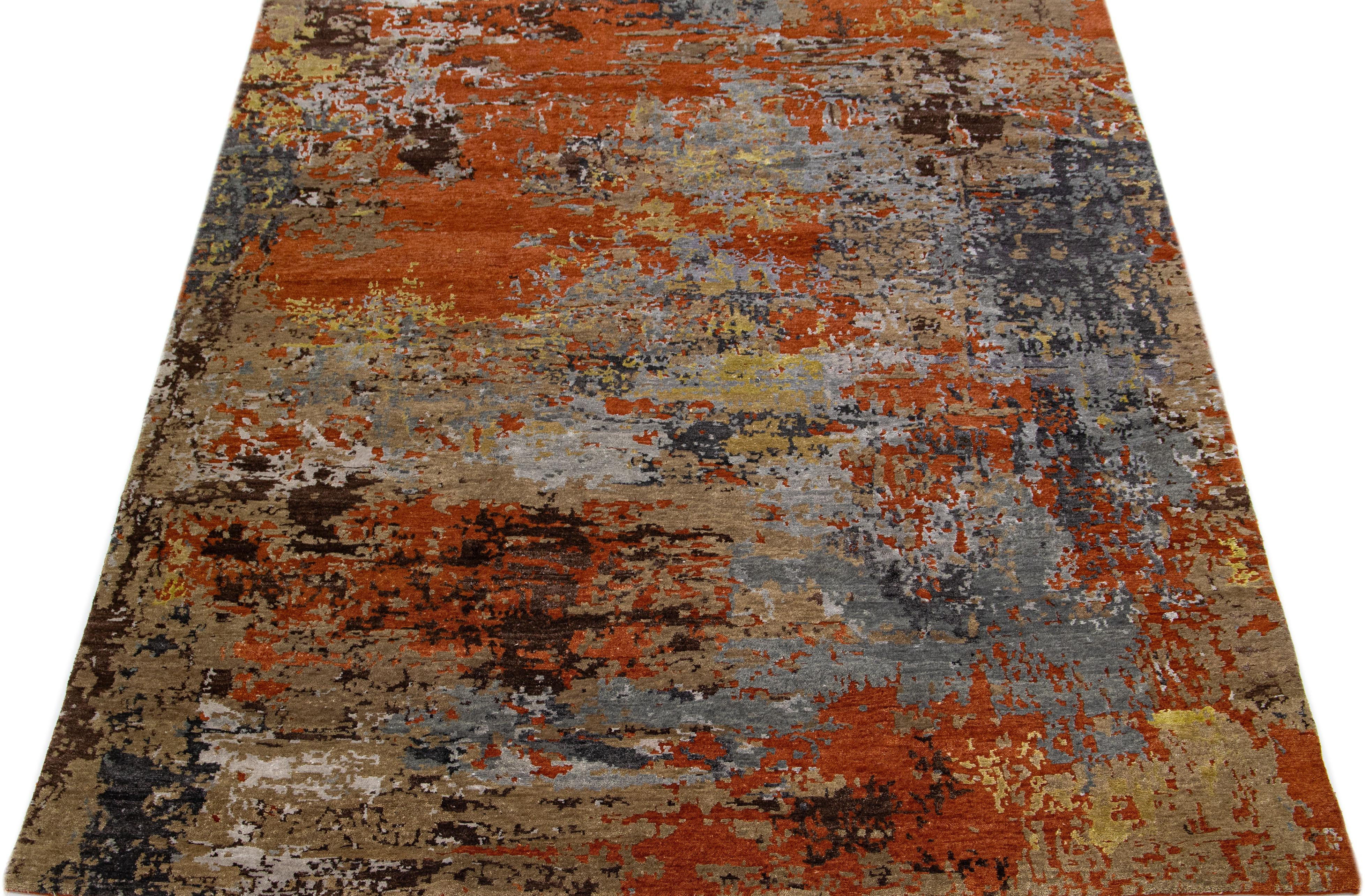 This Indian wool and silk blend rug features an orange field with an abstract pattern detailing gray and golden colors. Its composed materials provide robustness and longevity, while its ornamental design infuses any room with