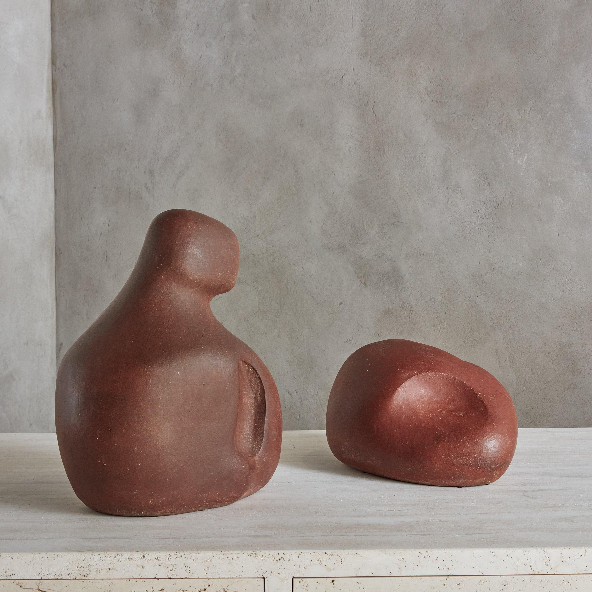 An Abstract, organic form terracotta clay sculpture featuring a smooth, red surface with natural aged patina. Delicately carved indents complement the elegant curves and rounded shape. The center of each sculpture is hollow with a hole present on