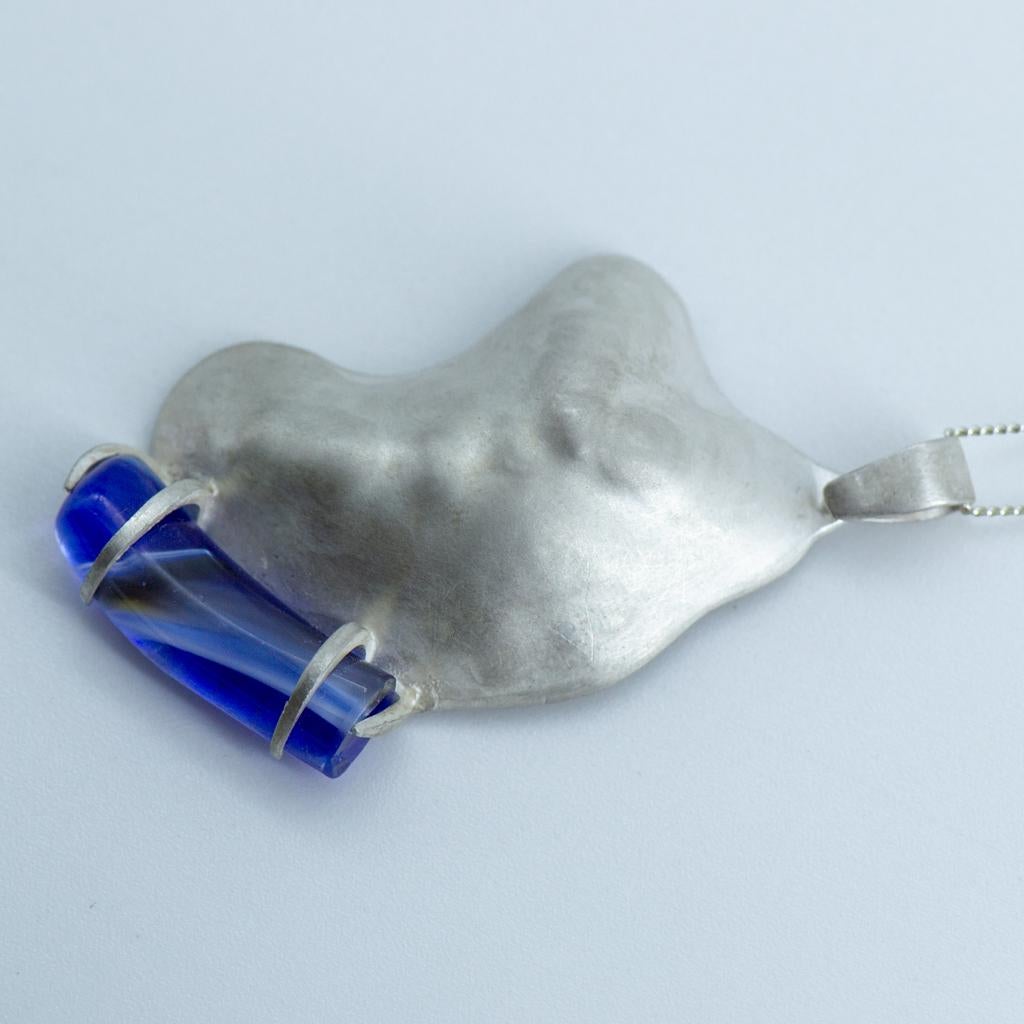 Questo ciondolo astratto contemporaneo in argento è stato cesellato a mano con le tradizionali tecniche di repoussé in una forma organica e fluida che rappresenta i contorni di una collina di Gozo, a Malta. Un frammento di vetro Mdina Glass di un