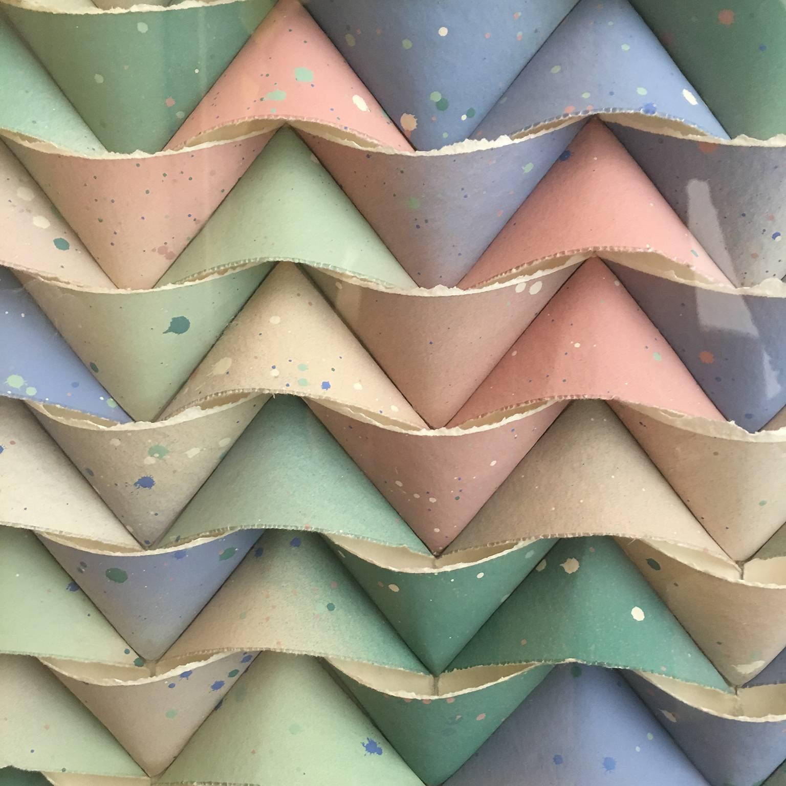 An original three dimensional paper collage by JoAnne Casey. This piece features raised geometric shapes of paper in subtle hues of blue, yellow, orange, red, green, purple, and grey. The artists signature can be found in the lower left of the