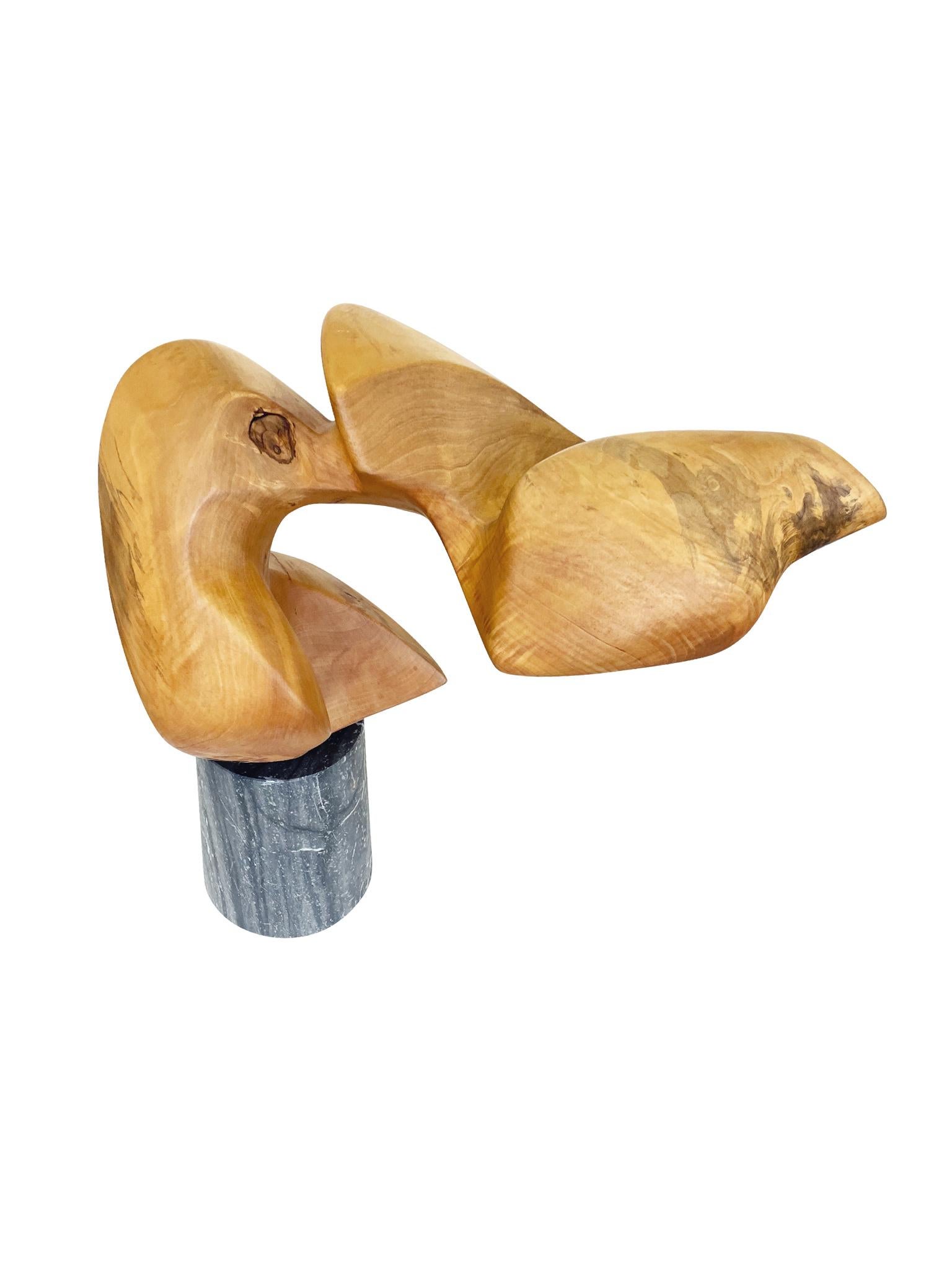 An abstract sculpture consisting of hand-carved wood with a gray marble base. The wood's biomorphic shape calls to mind a figure striking a dynamic pose. Bold grain patterning and a honey-yellow tone characterize the smooth surface of the wood. A