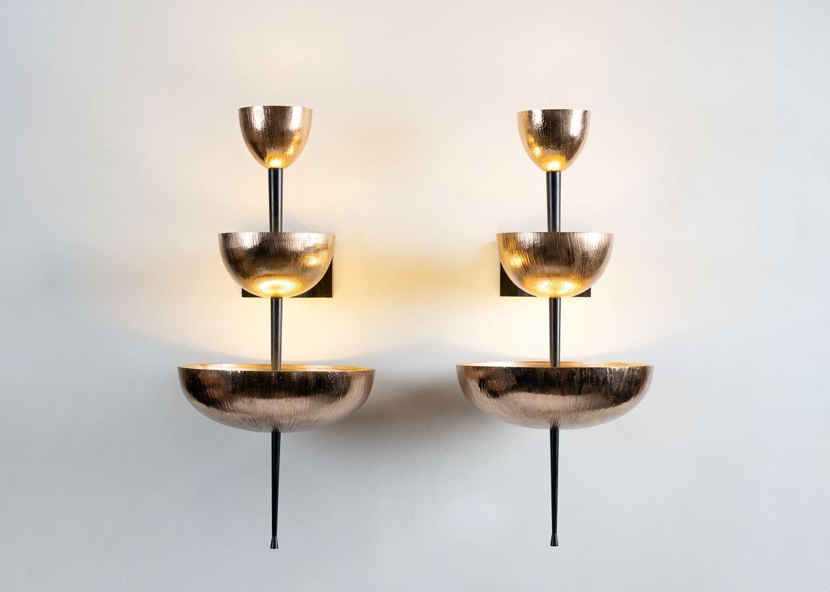 Simposio, a contemporary wall sconce by Roman architect Achille Salvagni, features a staggered succession of three vertically arranged, patinated bronze bowls bisected by a pointed, burnished bronze shaft. 

Limited edition of 20 + AP.