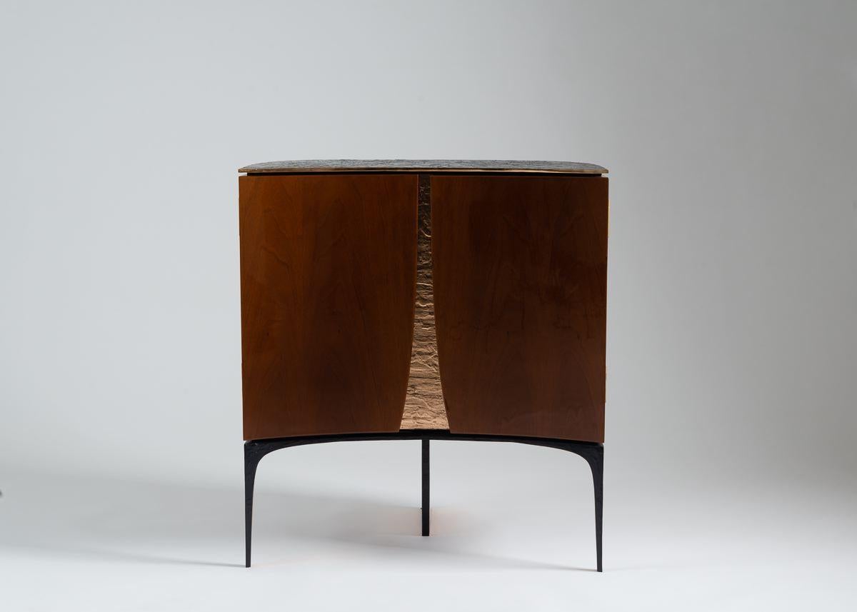 Achille Salvagni's beautiful bedside tables have sensually concave, double doors of sleek walnut separated by the glimmer of warm, bright, hand-hammered bronze. Each rests on three legs, the central one of which is, itself, a visual blade of
