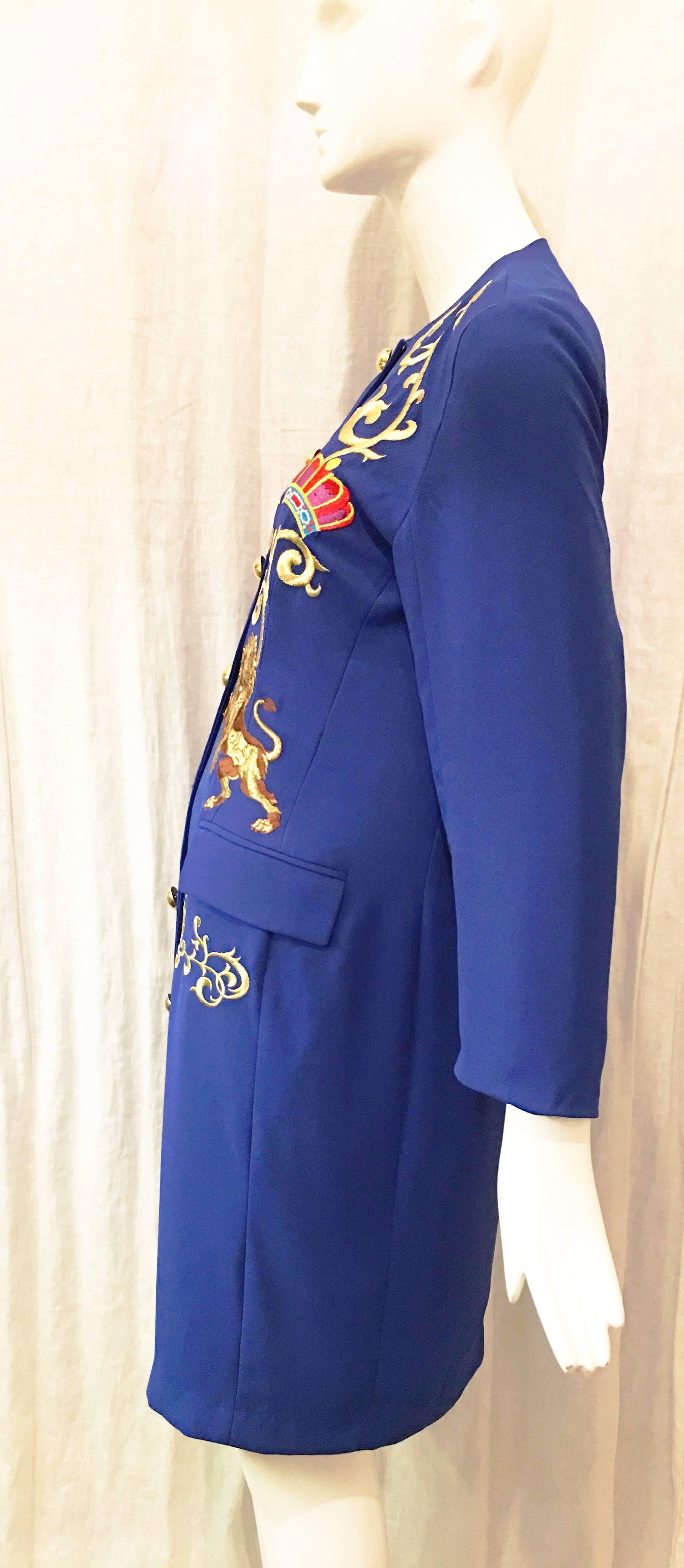 Cobalt blue long blazer-style jacket with embroidered lions. Seven gold buttons down the front of the jacket (one button missing). Gold tone curly cue embroidered design above crown and lion embroidery and at front waist. Collarless. Sleeves hit