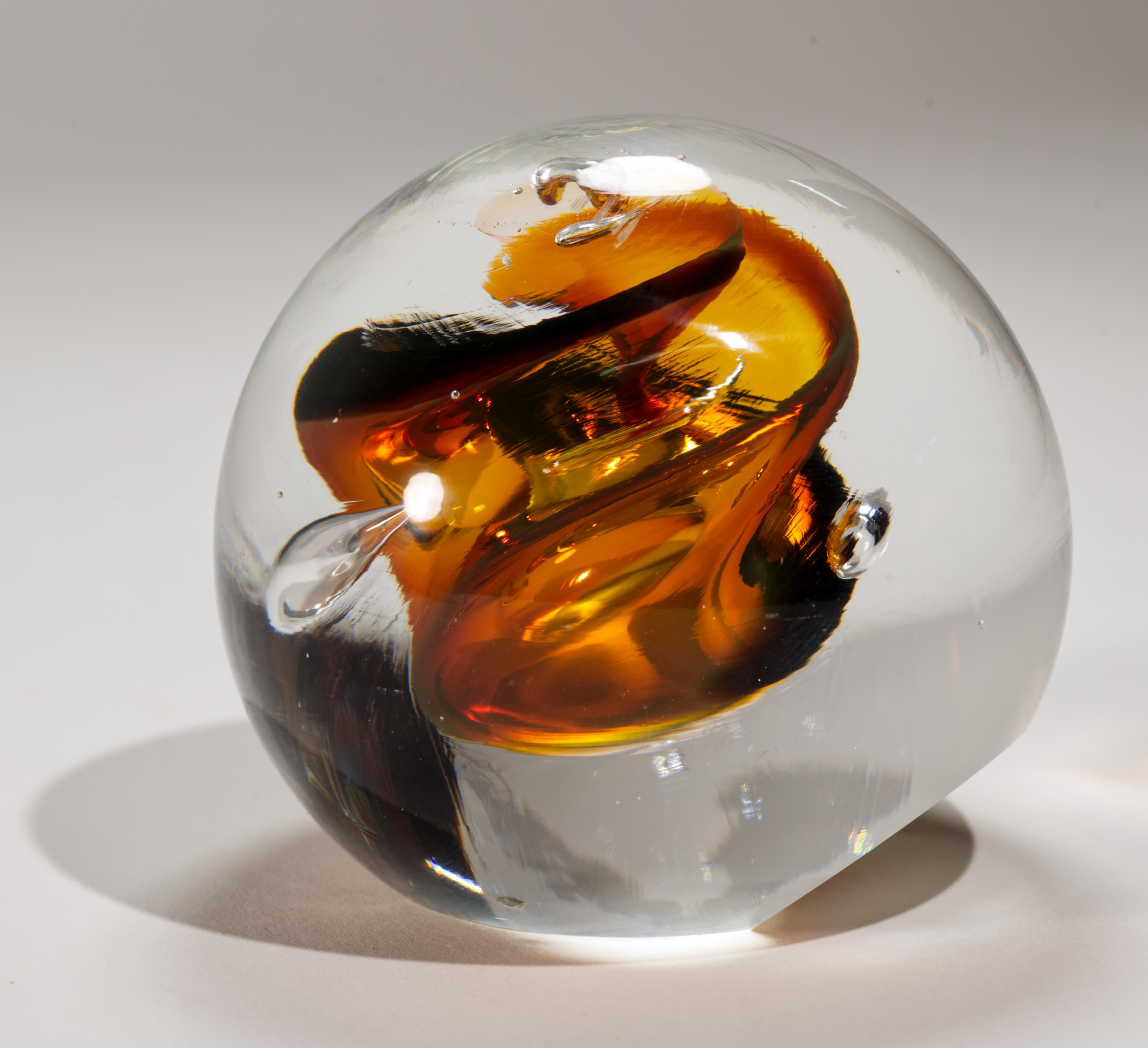 Striking abstract art glass paperweight features central core of swirling deep amber, cognac brown, and opal colored glass with controlled bubbles, enclosed in a layer of clear glass in sommerso technique. Rounded paperweight has slanted base that