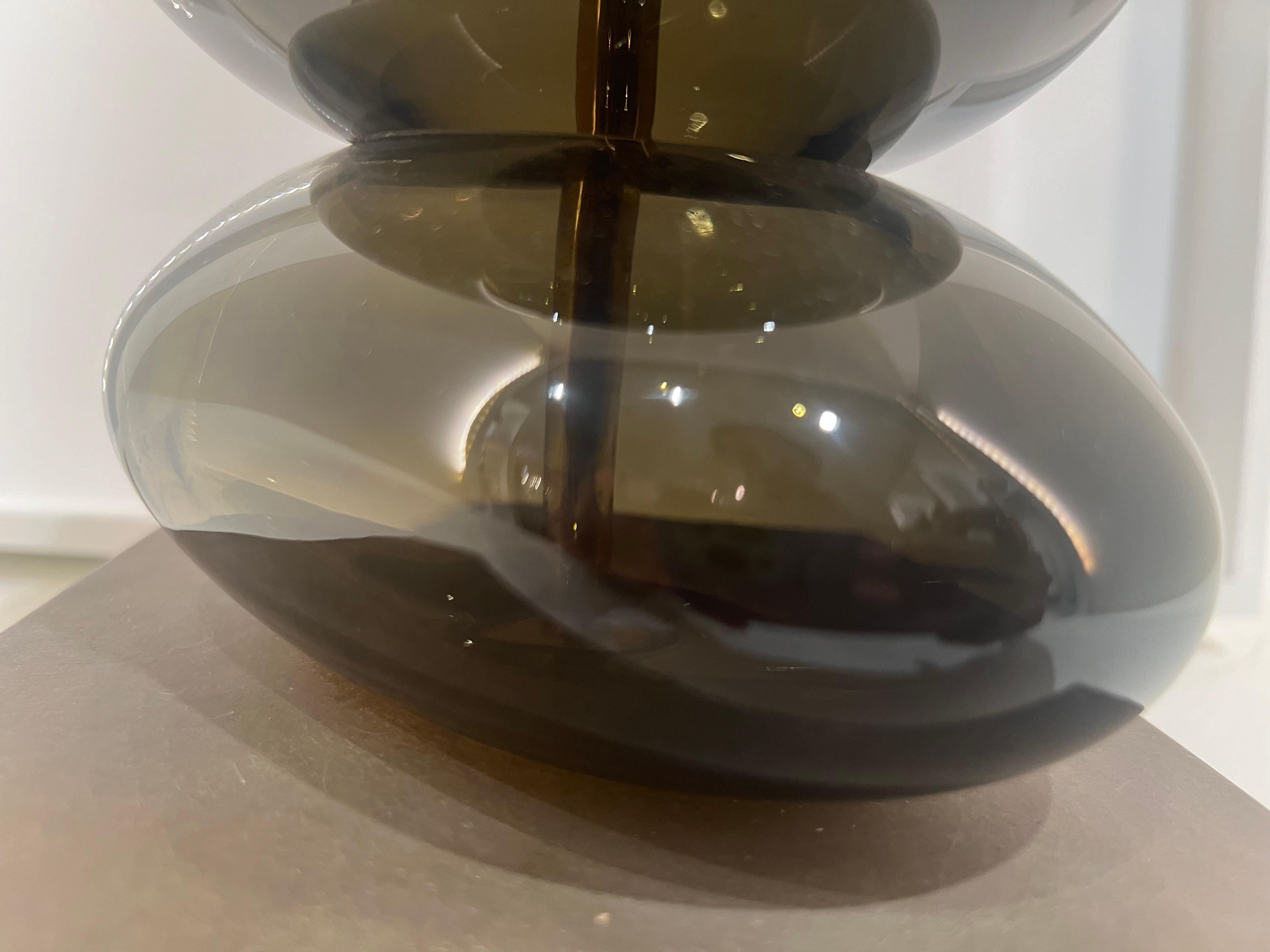 The Adam Lamp was designed by British glass artist Adam Aaronson. Resembling a stack of pebbles on the beach, each glass ‘pebble’ is blown, ground and polished to balance perfectly in charcoal glass with Antique Brass base.

