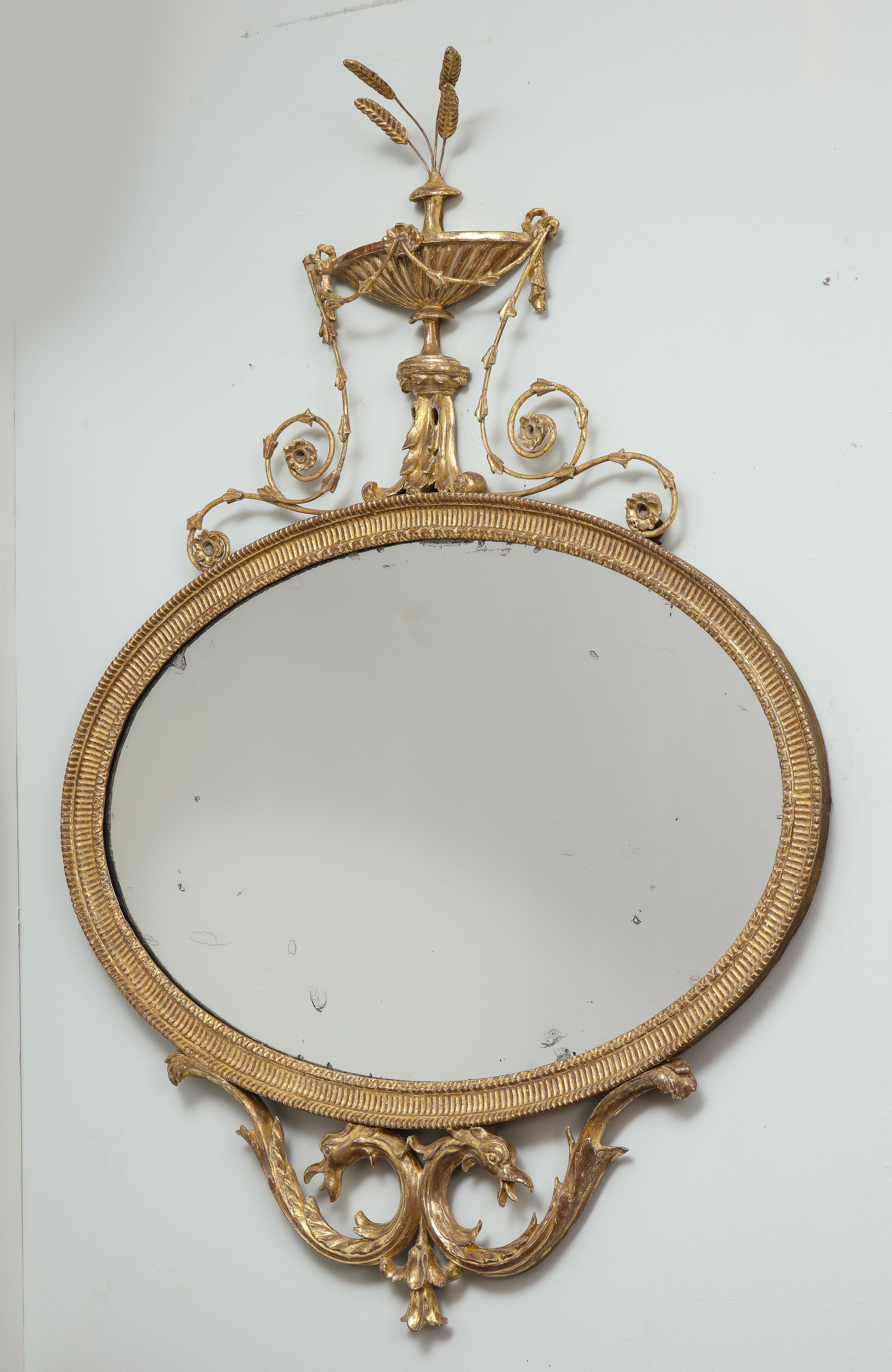 Fine George III period giltwood mirror, the classical urn finial with sprouting wheat ears, the urn draped with garlands of bell flowers surmounting the ribbed and lamb tongue carved oval frame having original speckled mercury glass plate, the base
