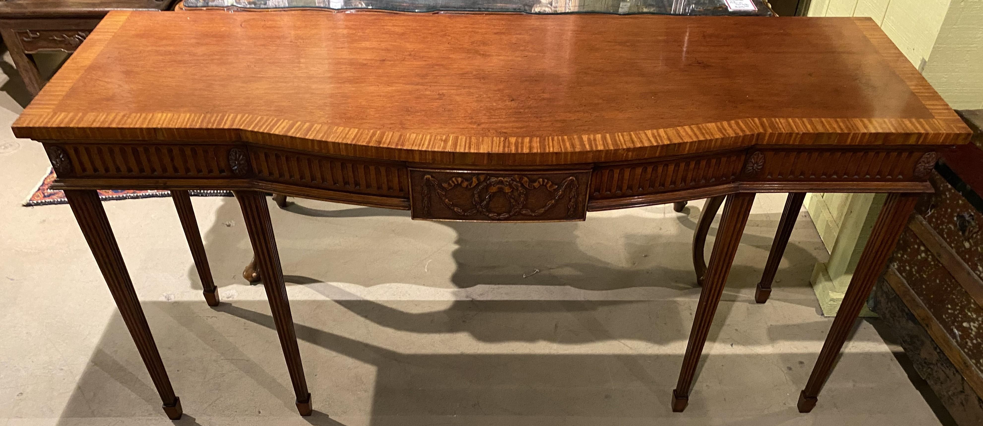 A fine example of an Adam style English mahogany serpentine front server or sideboard with conforming top with crossbanded veneer border surmounting a single fitted frieze drawer with center carved wreath/swag cartouche, carved foliate corner