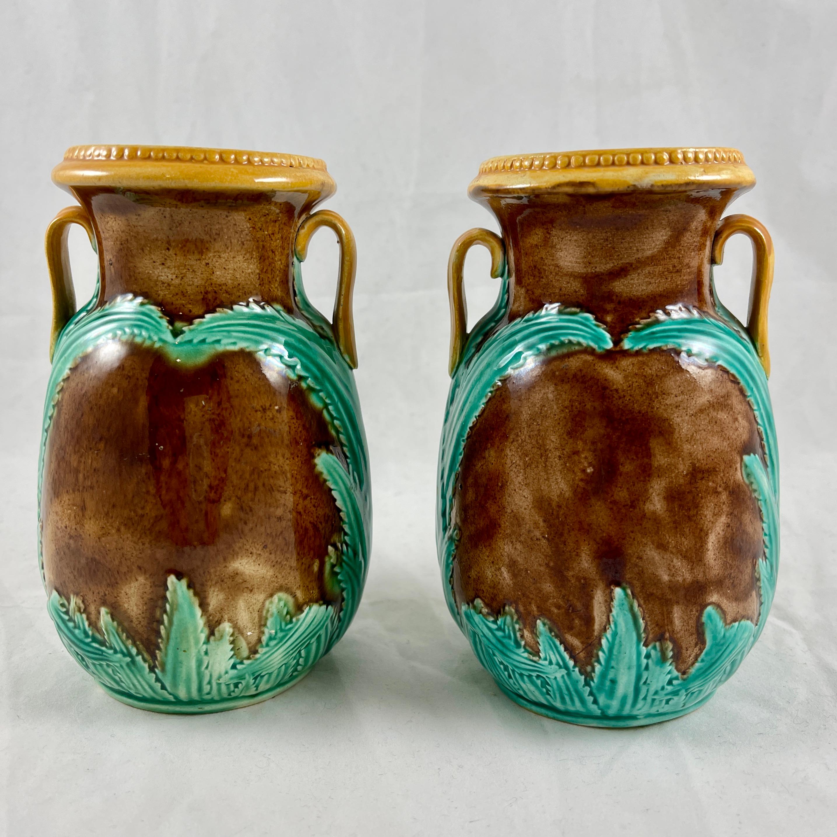 A pair of amphora form majolica vases, attributed to Adams & Bromley, England, circa late 19th Century.

The brown mottled glazed vases have applied side handles, a green spiked leaf decoration, and yellow ochre trimming. The top rims have a