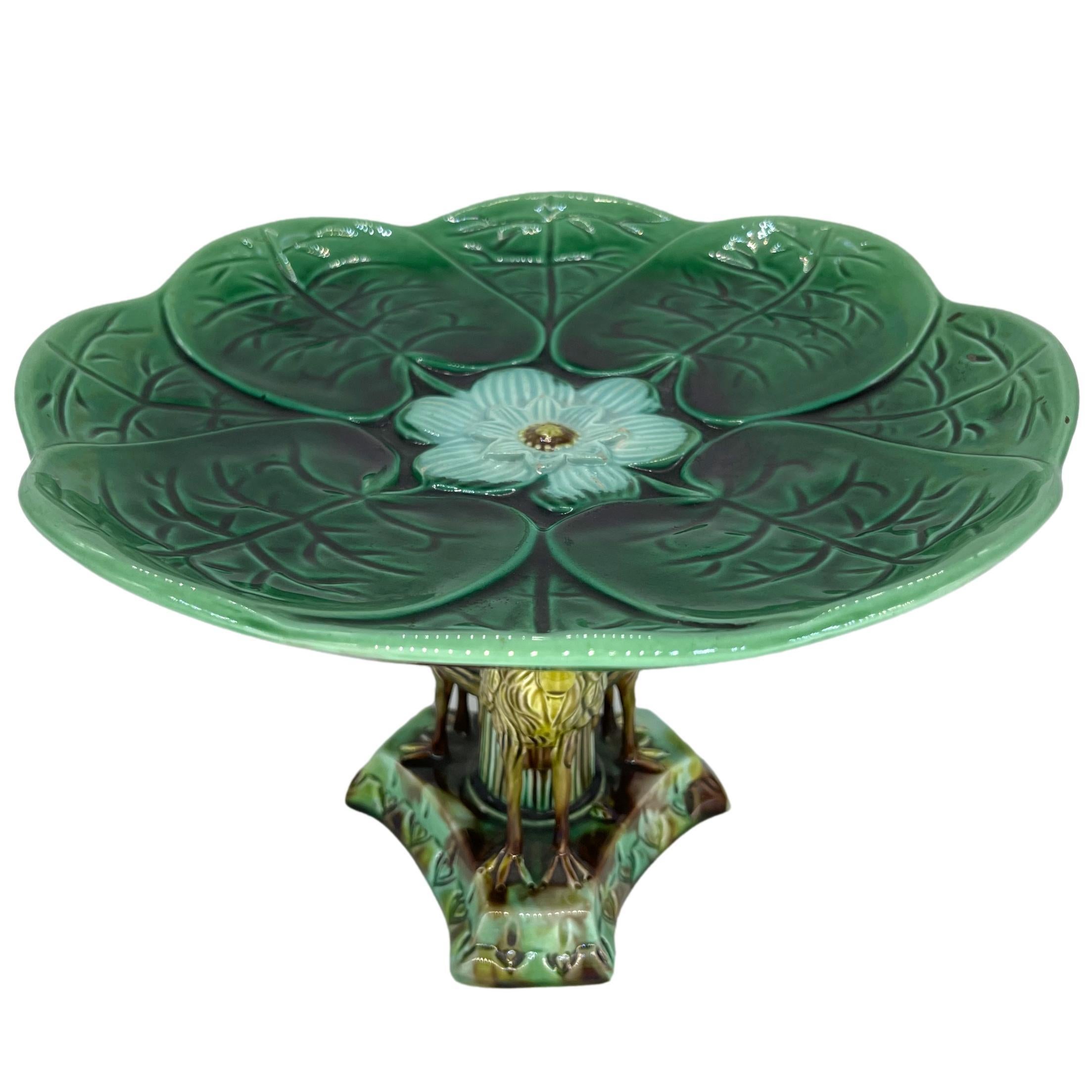 Adams & Bromley Majolica Comport Design Number 1, the dish formed by green-glazed lily pads with a central relief molded blossom, supported by a stalk-form column with three applied herons, on a raised plinth pedestal base, English, ca. 1880. 
For