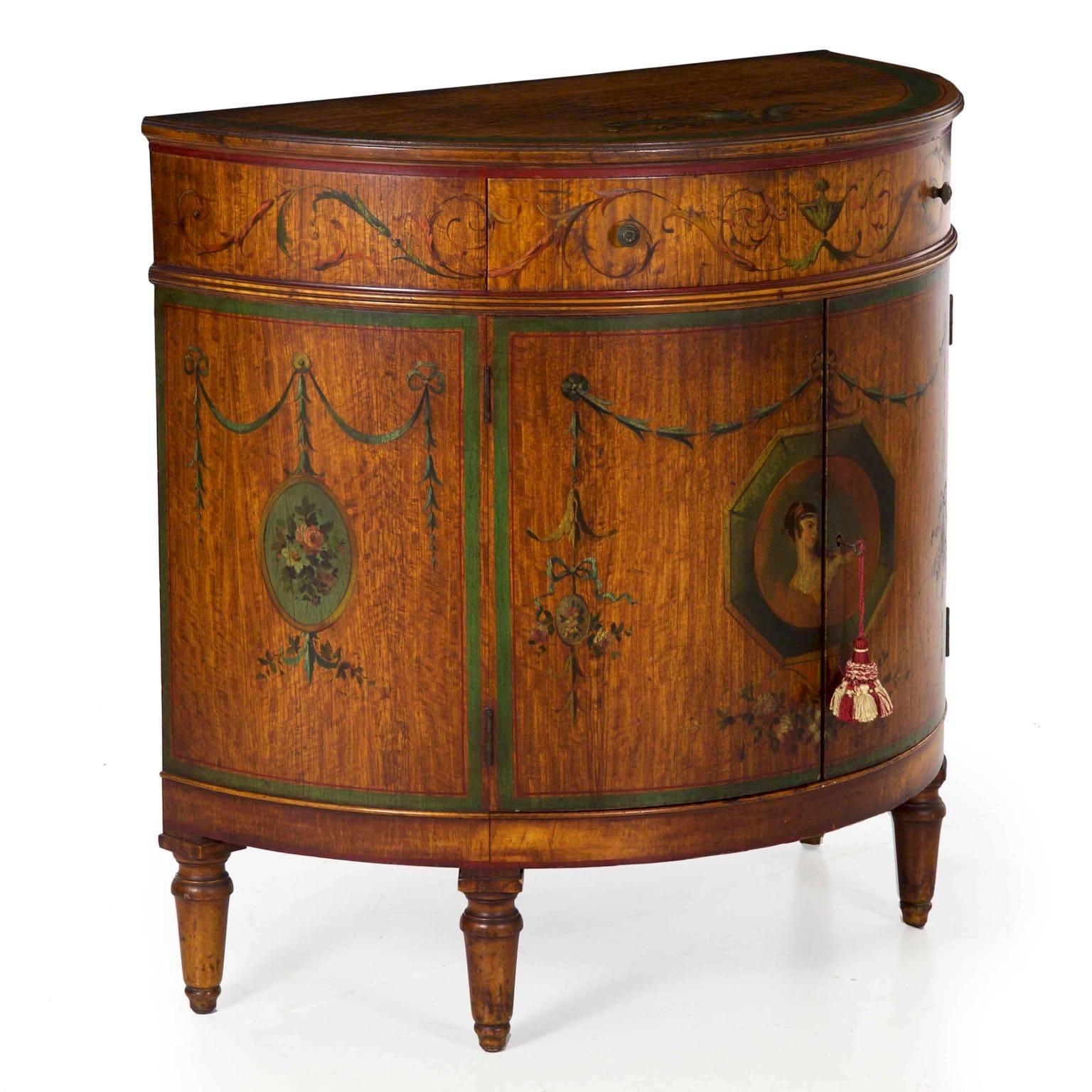 This is an incredibly striking demilune cabinet with exquisite painted surface detailing throughout. The garlands of draped foliage, the urn in the centre of the drawer, the strings of honeysuckle and flowing ribbon, everything is expertly painted
