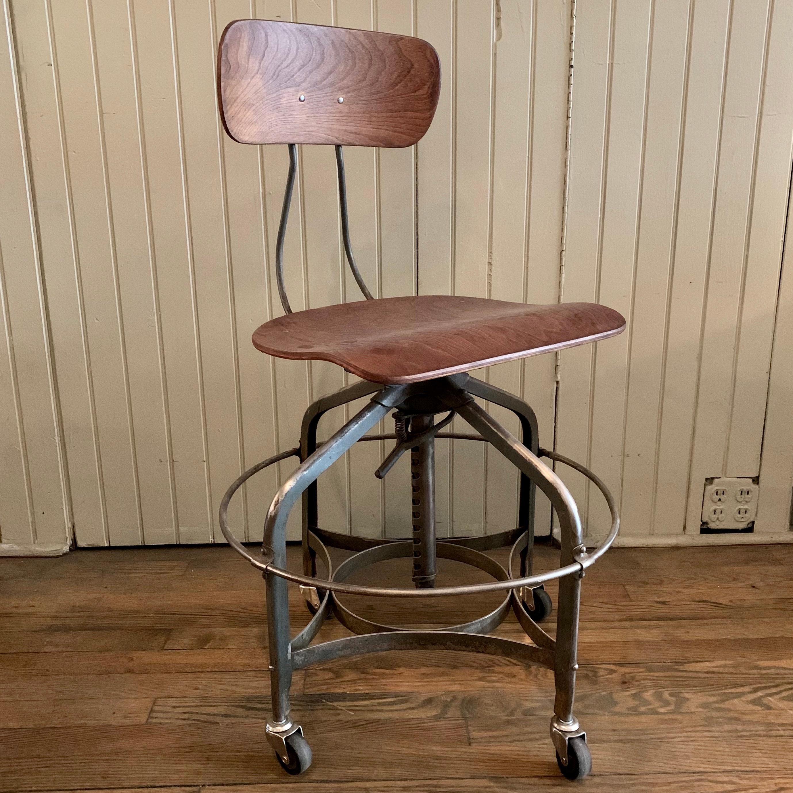 Industrial, adjustable, rolling, factory stool by Toledo Metal Furniture Co., features a brushed steel frame with wide 20 inch diameter ring footrest and oak seat and back. The height adjusts from 20 - 28 inches. The seat measures 16 wide x 14 deep.