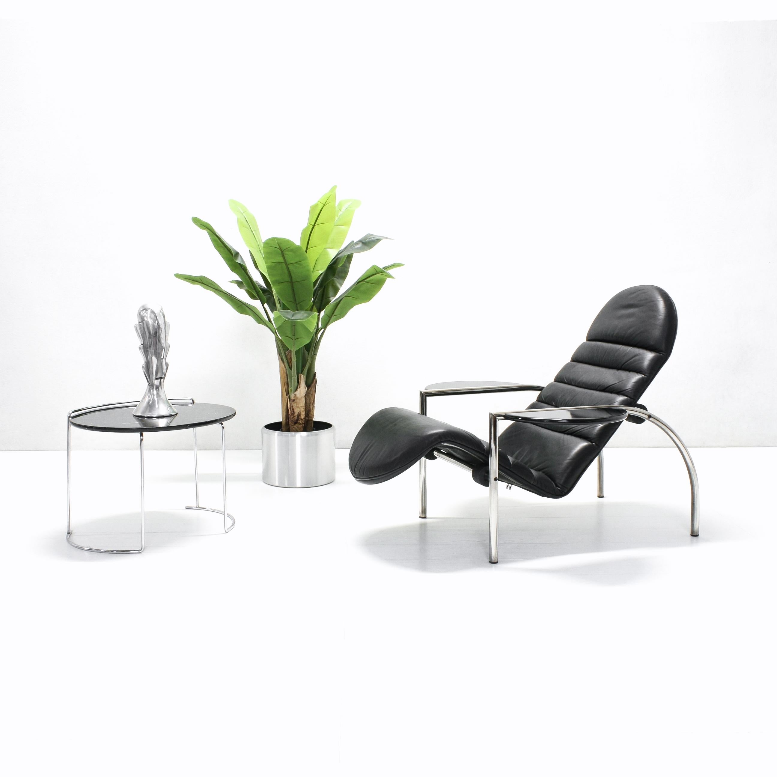 This model Noe chair features a chromed metal tube frame with a black leather seat and black lacquered wooden armrests that can function easily as tables. Thanks to a simple, yet functional roller system, the chair can be used for both sitting as